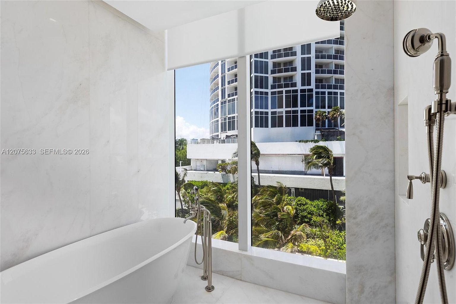 This luxurious primary bathroom features a sleek, freestanding soaking tub positioned next to large windows that offer a view of the surrounding tropical landscape and high-rise buildings. The space is finished with light-toned stone wall cladding and includes a modern shower fixture with a handheld sprayer and overhead rainfall head. The overall aesthetic is bright, clean, and sophisticated, emphasizing a spa-like atmosphere.