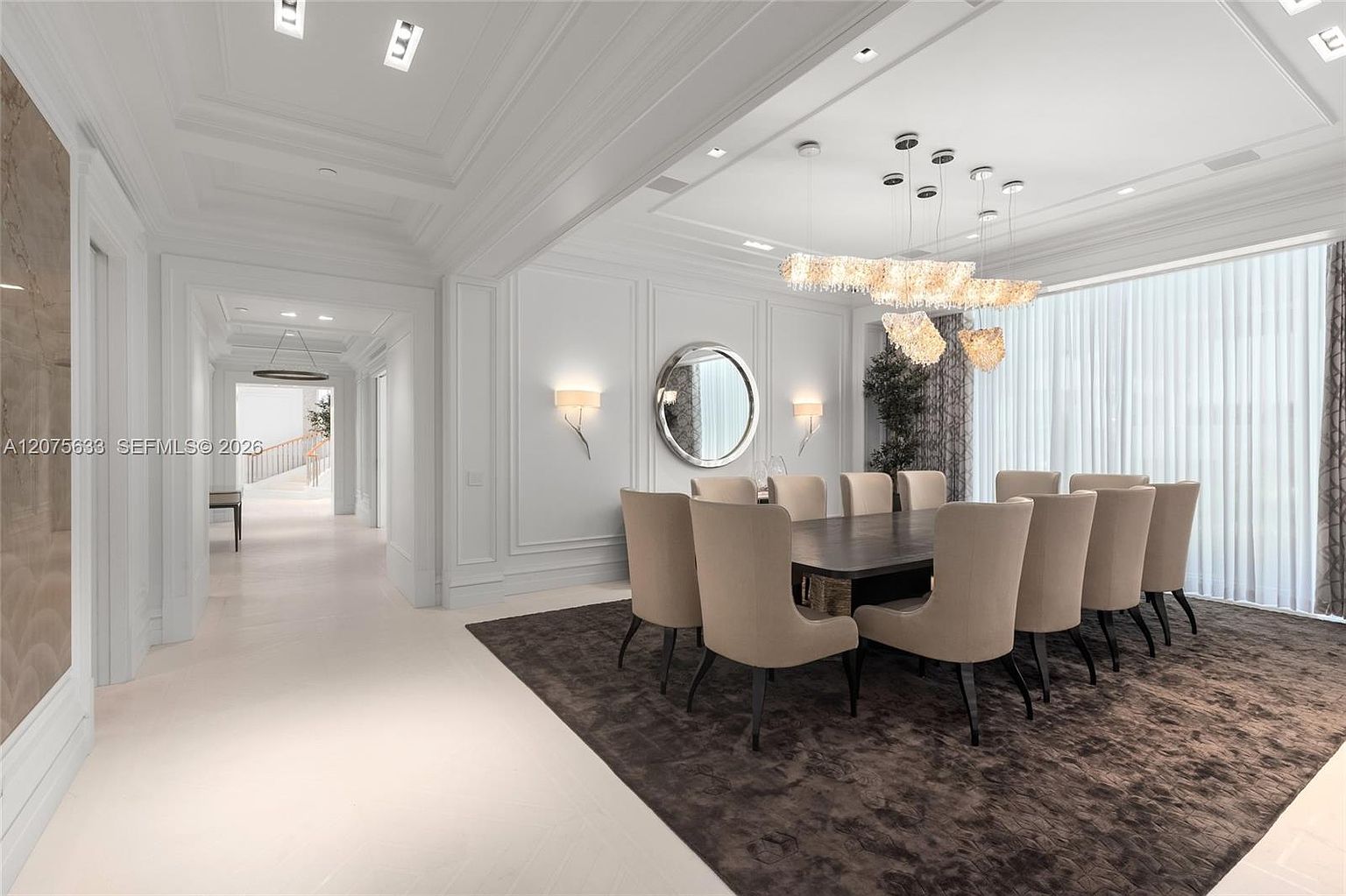 This elegant dining room features a large, dark-toned rectangular table surrounded by ten upholstered chairs in a neutral beige fabric. A striking, multi-tiered crystal chandelier serves as the focal point above the table, while a circular mirror and wall sconces add a sophisticated touch to the white-paneled walls. The space is grounded by a large, dark textured area rug and features expansive windows covered by sheer floor-to-ceiling drapery, creating a bright and luxurious atmosphere.
