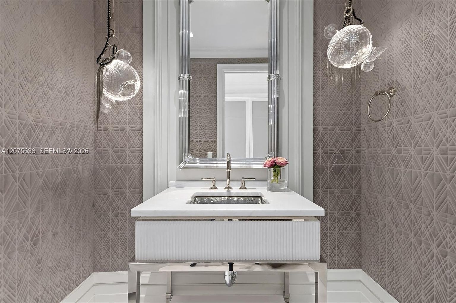This elegant powder room features a sleek, wall-mounted vanity with a white stone countertop and a metallic basin. The walls are adorned with a sophisticated geometric patterned wallpaper in a neutral tone, complemented by unique, organic-shaped glass pendant lights. A tall, framed mirror with fluted side columns serves as the focal point, creating a refined and luxurious atmosphere.