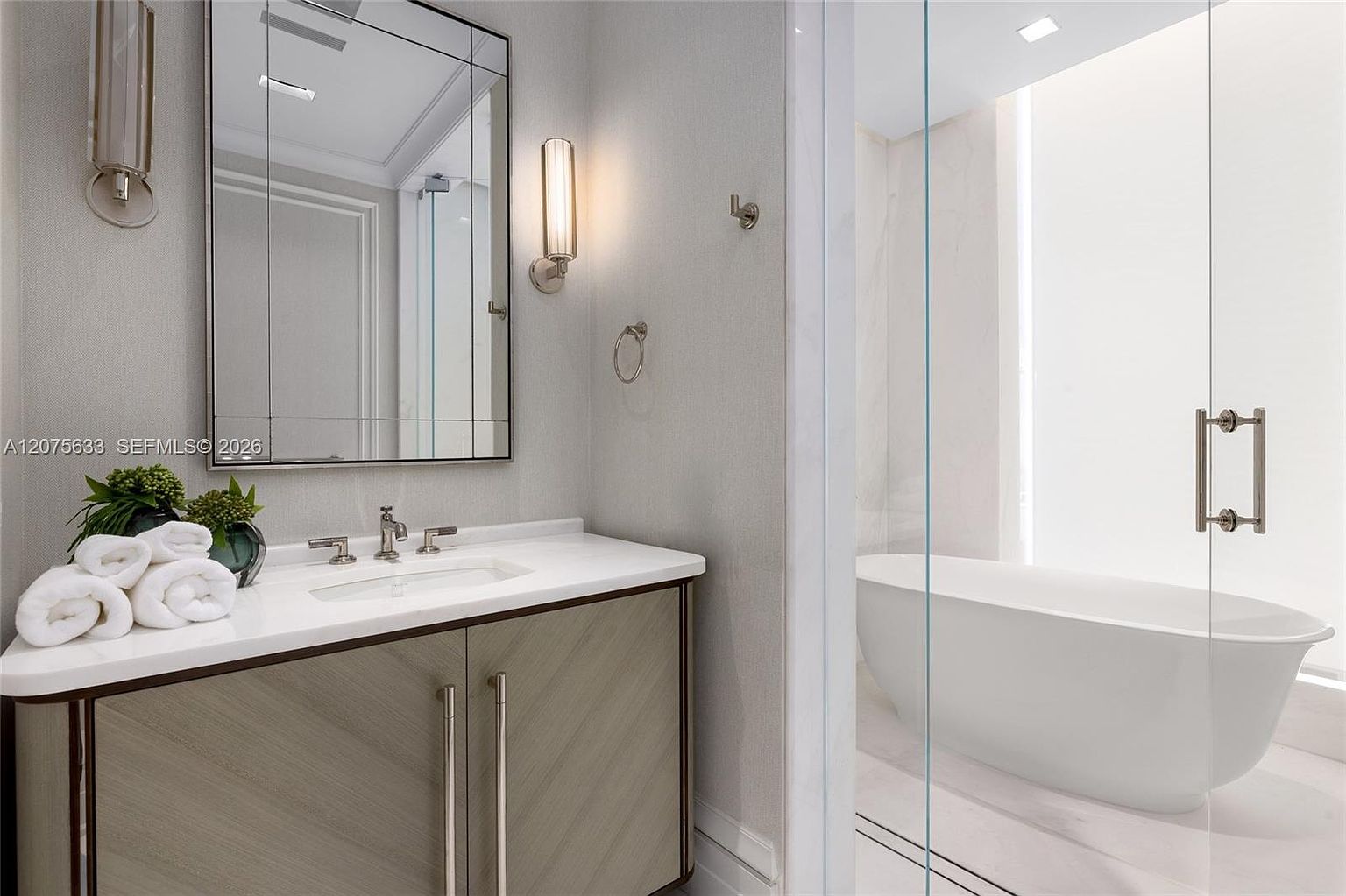 This modern bathroom features a sleek vanity with a light-toned, chevron-patterned cabinet and a crisp white countertop. A rectangular mirror is flanked by two vertical wall sconces, while a glass partition separates the vanity area from a bright, airy space containing a freestanding soaking tub. The room is finished in neutral tones, creating a clean, sophisticated, and spa-like atmosphere.