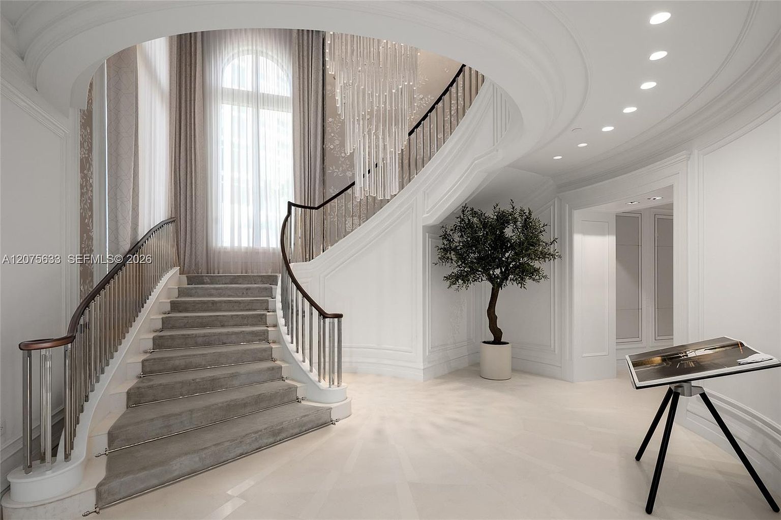 This grand foyer features a sweeping curved staircase with a dark wood handrail and metal balusters, leading up to a landing. A dramatic, multi-tiered crystal chandelier hangs from the ceiling, casting a sophisticated glow over the space, while a potted tree adds a touch of greenery near the entryway. The area is finished with light-toned flooring and crisp white walls, creating an elegant and airy atmosphere.