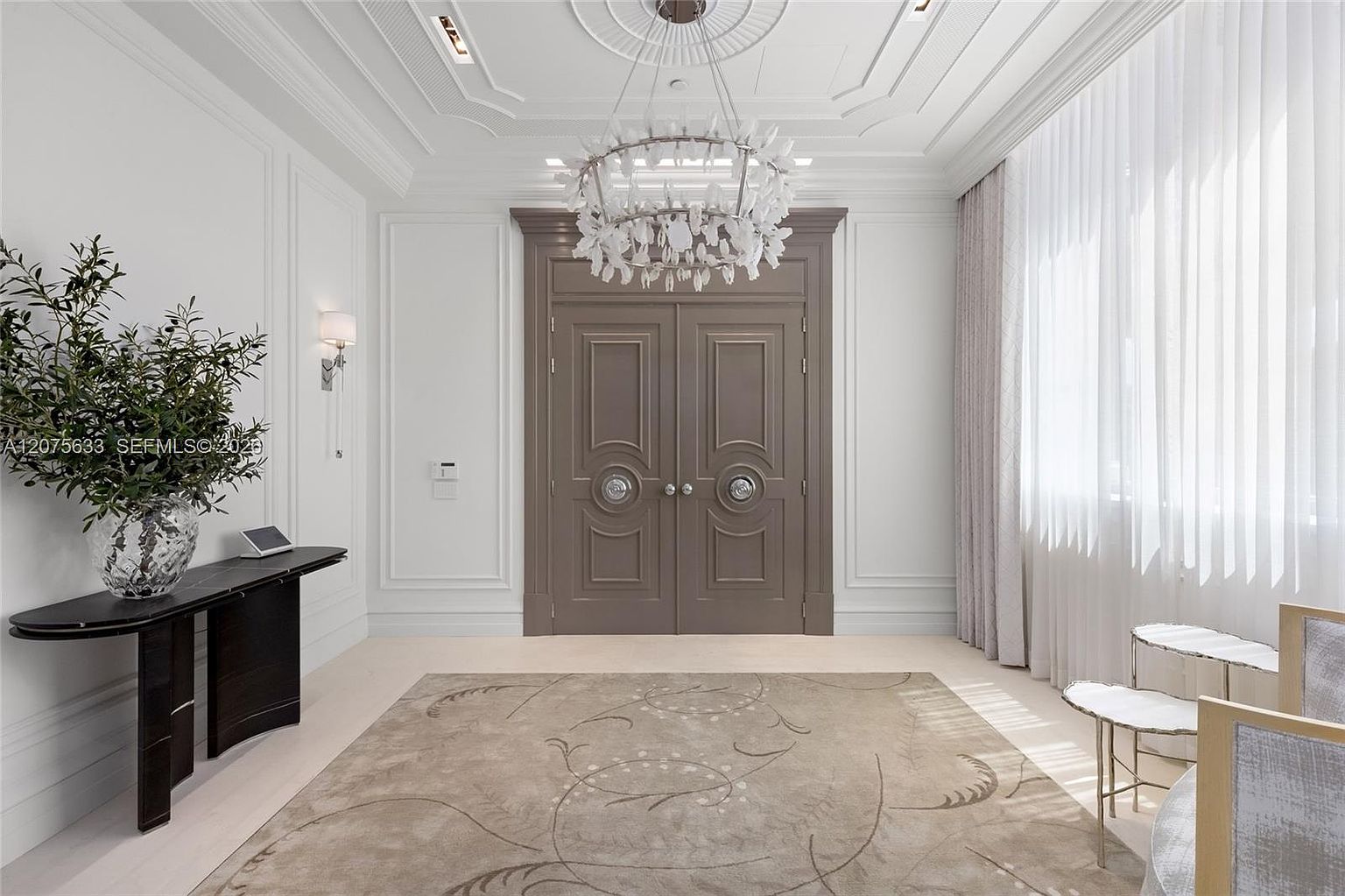 This elegant foyer features a grand set of double doors with circular hardware, serving as the focal point of the space. A large, intricate chandelier hangs from a detailed ceiling medallion, casting light over a neutral-toned area rug and a sleek, dark console table adorned with a decorative plant. The room is framed by crisp white walls with classic molding and tall, sheer curtains that soften the natural light entering from the side.