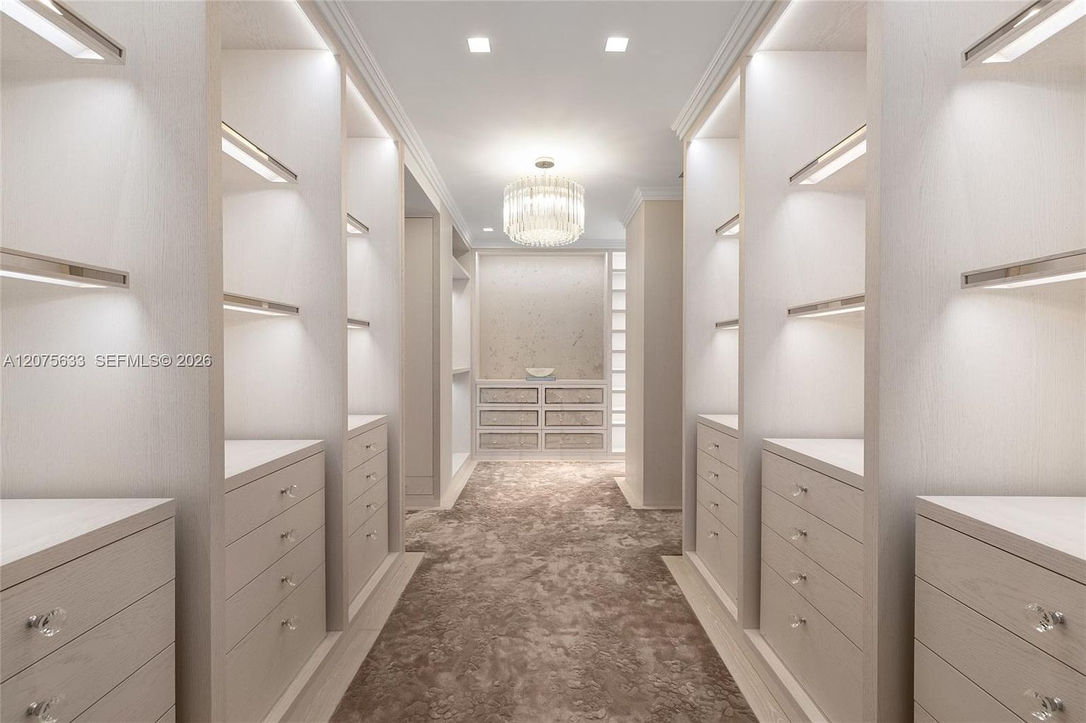 This luxurious walk-in closet features symmetrical light wood cabinetry with integrated LED shelf lighting and crystal-style hardware. A central crystal chandelier hangs from the ceiling, casting a warm glow over the textured, neutral-toned carpet. The space is designed with a clean, sophisticated aesthetic, offering ample storage and a refined, boutique-like atmosphere.