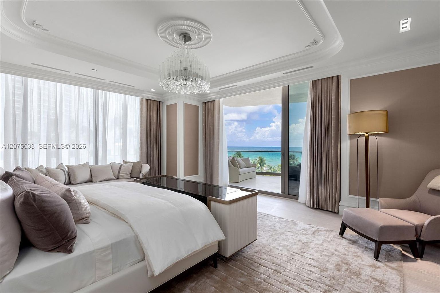 This luxurious primary bedroom features a neutral color palette with soft beige and cream tones, centered around a large bed with a dark wood footboard console. The room is illuminated by a crystal chandelier and a tall floor lamp, while floor-to-ceiling glass doors open onto a private balcony with ocean views. A plush area rug and a comfortable armchair create a serene, high-end atmosphere.
