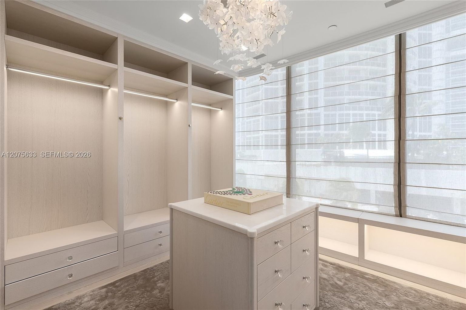 This sophisticated walk-in closet features custom light-toned cabinetry with integrated LED lighting and a central island topped with a stone surface. A large, intricate chandelier hangs from the ceiling, while expansive windows with light-filtering shades provide a bright, airy atmosphere. The space is finished with a neutral-toned carpet, creating a luxurious and organized dressing area.