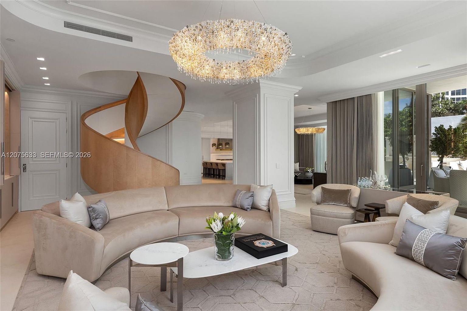 This elegant, open-concept living room features a large, curved sectional sofa arranged around a modern coffee table, creating a sophisticated seating area. A striking, circular crystal chandelier serves as the focal point, suspended above the space, while a sculptural, light-toned wood staircase curves gracefully in the background. The room is bathed in natural light from large windows and maintains a neutral, luxurious color palette that flows into an adjacent kitchen area.