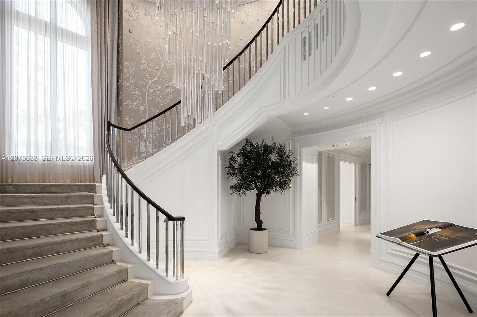 This grand foyer features a sweeping curved staircase with a dark wood handrail and metal balusters, leading to an upper level. A dramatic crystal chandelier hangs from the ceiling, casting light over the space, while a potted tree adds a touch of greenery against the white walls. The area is bright and airy, with large windows draped in floor-to-ceiling curtains and a minimalist table displaying a book in the foreground.