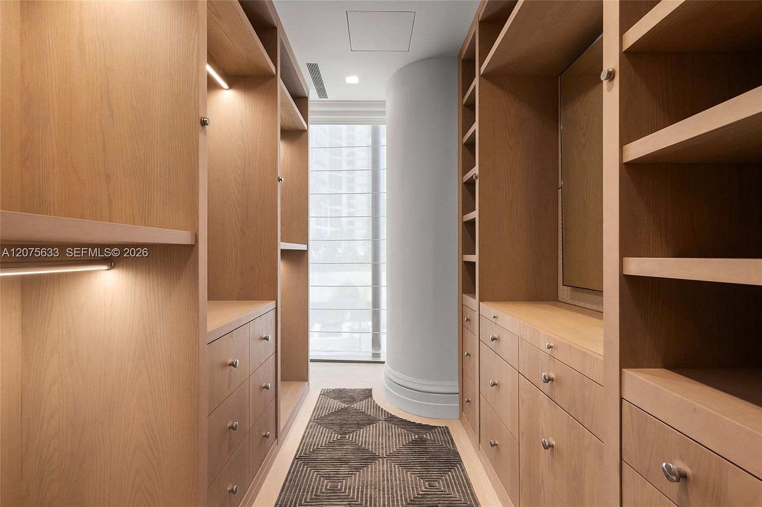 This walk-in closet features custom light wood cabinetry with integrated LED lighting, providing ample storage and a warm, organized aesthetic. A geometric patterned runner rug centers the space, leading toward a window with blinds that allows natural light to filter in. The layout includes a mix of open shelving, hanging rods, and multiple drawers, creating a functional and sophisticated dressing area.