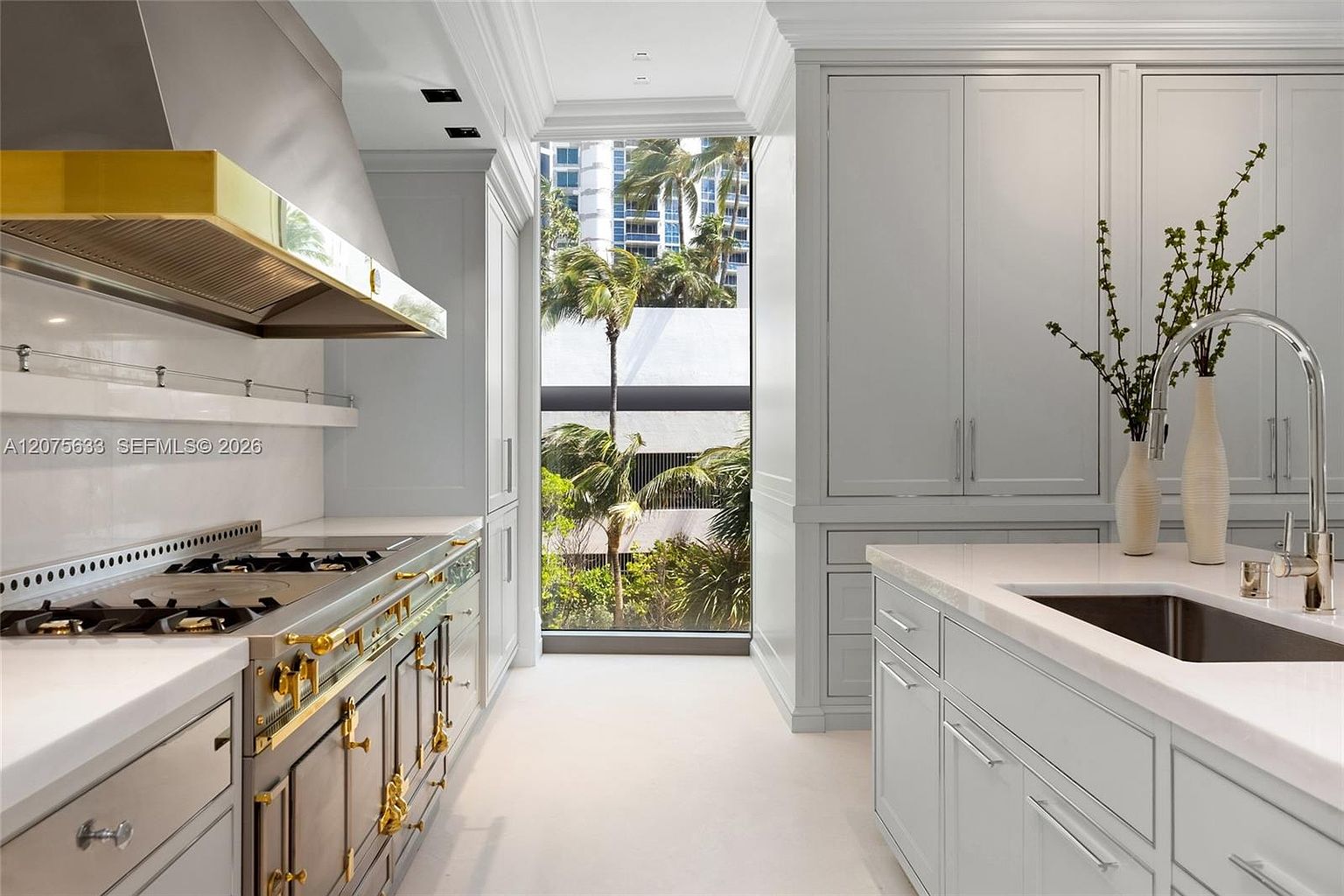 This elegant, high-end kitchen features light-toned cabinetry, a professional-grade range with a prominent gold-toned hood, and sleek stone countertops. A large, floor-to-ceiling window at the end of the galley layout provides a bright, natural view of palm trees and the exterior surroundings. The space is designed with a clean, sophisticated aesthetic, balancing modern luxury with functional workspace.