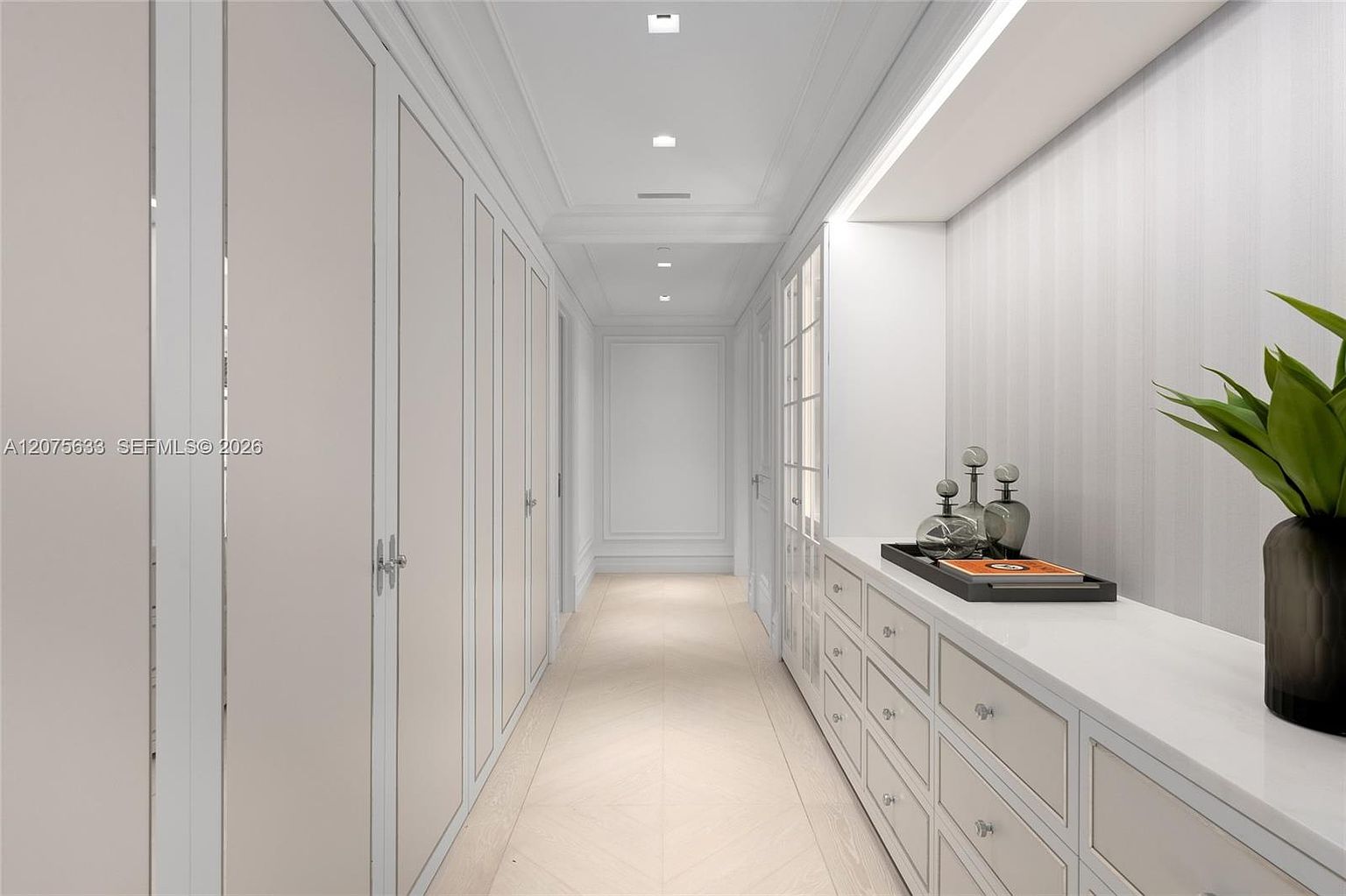 This elegant, light-filled walk-in closet features a long, symmetrical hallway lined with floor-to-ceiling cabinetry and integrated lighting. The space is finished in a neutral, monochromatic palette with light-toned wood flooring and clean, minimalist lines. A built-in dresser unit on the right provides surface space for decor, while recessed ceiling lights create a bright and sophisticated atmosphere.