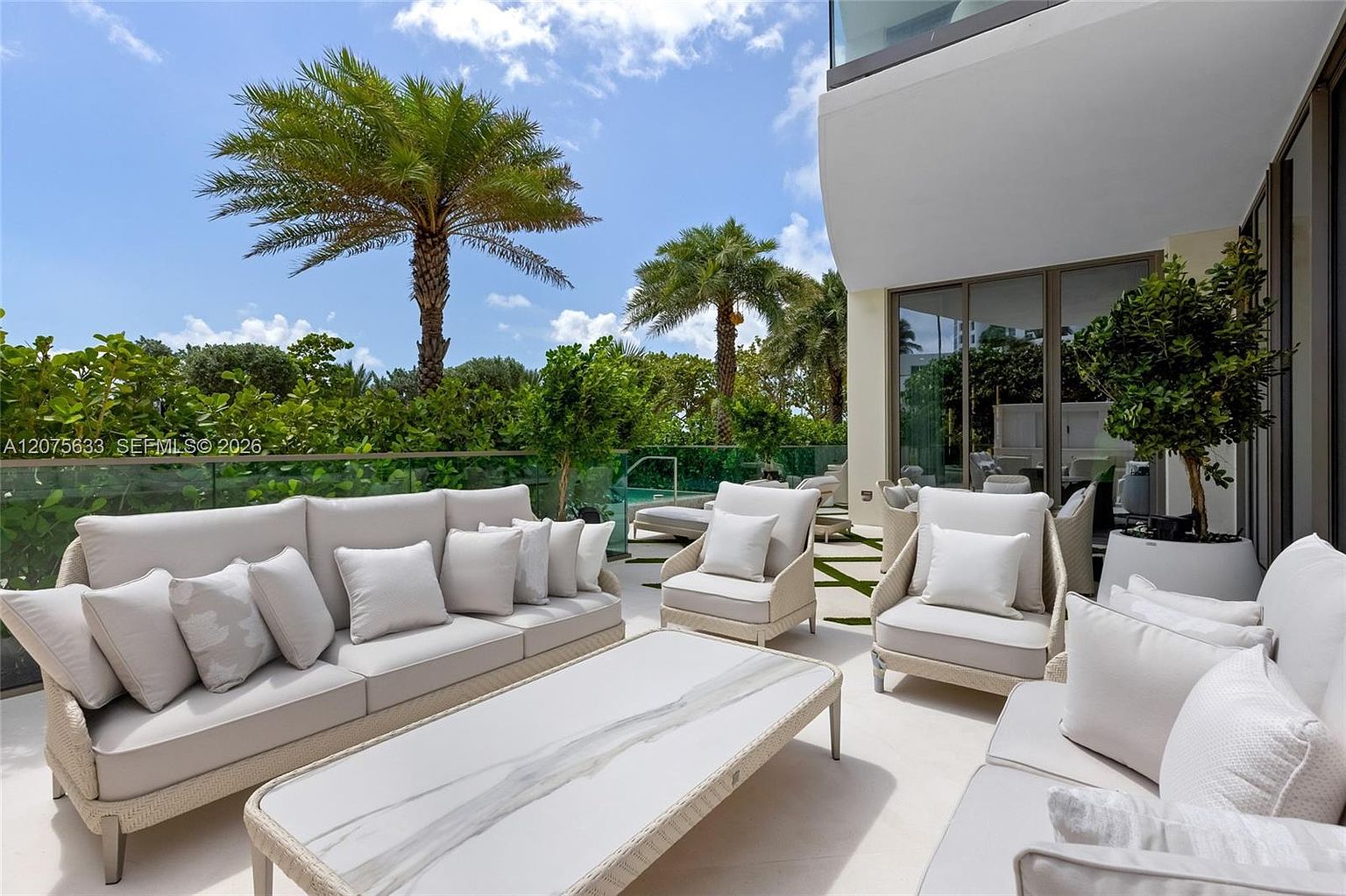 This expansive, sun-drenched terrace features a sophisticated outdoor lounge area furnished with plush, light-toned seating and a central stone-topped coffee table. The space is framed by a glass railing that offers an unobstructed view of lush tropical landscaping and a glimpse of a private pool. The modern architectural design seamlessly blends indoor and outdoor living, creating an inviting and luxurious atmosphere for relaxation.
