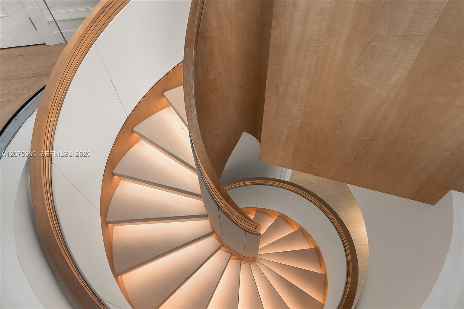 This high-angle, top-down perspective captures a striking spiral staircase featuring light wood treads and a curved, light wood handrail. The design is accented by integrated under-step lighting that casts a warm glow, contrasting with the clean, white curved walls. The architectural composition emphasizes the elegant, sweeping geometry of the staircase as it descends.