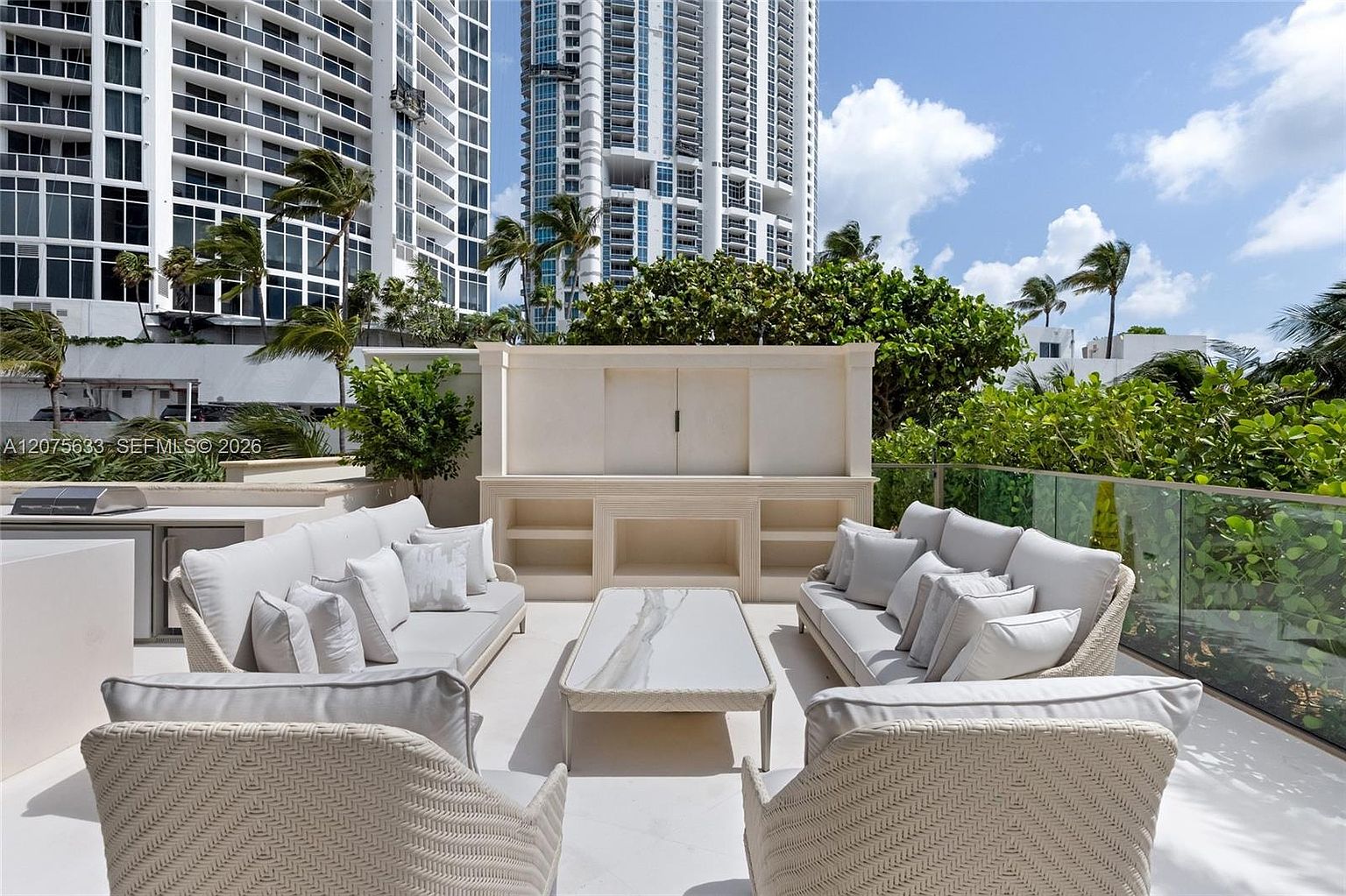 This expansive outdoor terrace features a sophisticated lounge area with two large, light-toned sofas facing each other around a central stone-topped coffee table. A custom-built, light-colored cabinetry unit with open shelving serves as a focal point against a backdrop of lush greenery and towering high-rise buildings. The space is enclosed by a sleek glass railing, offering a bright, airy, and luxurious atmosphere perfect for outdoor entertaining.