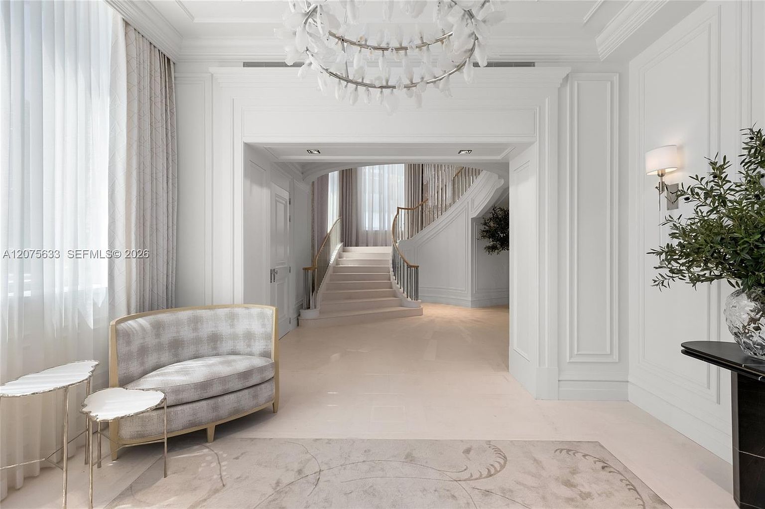 This elegant foyer features a grand, curved staircase as its focal point, framed by a wide, arched entryway. The space is decorated in a sophisticated, monochromatic palette with a patterned settee, delicate side tables, and a striking sculptural chandelier. The perspective is from the entryway looking toward the staircase, emphasizing the open, airy, and luxurious architectural design.