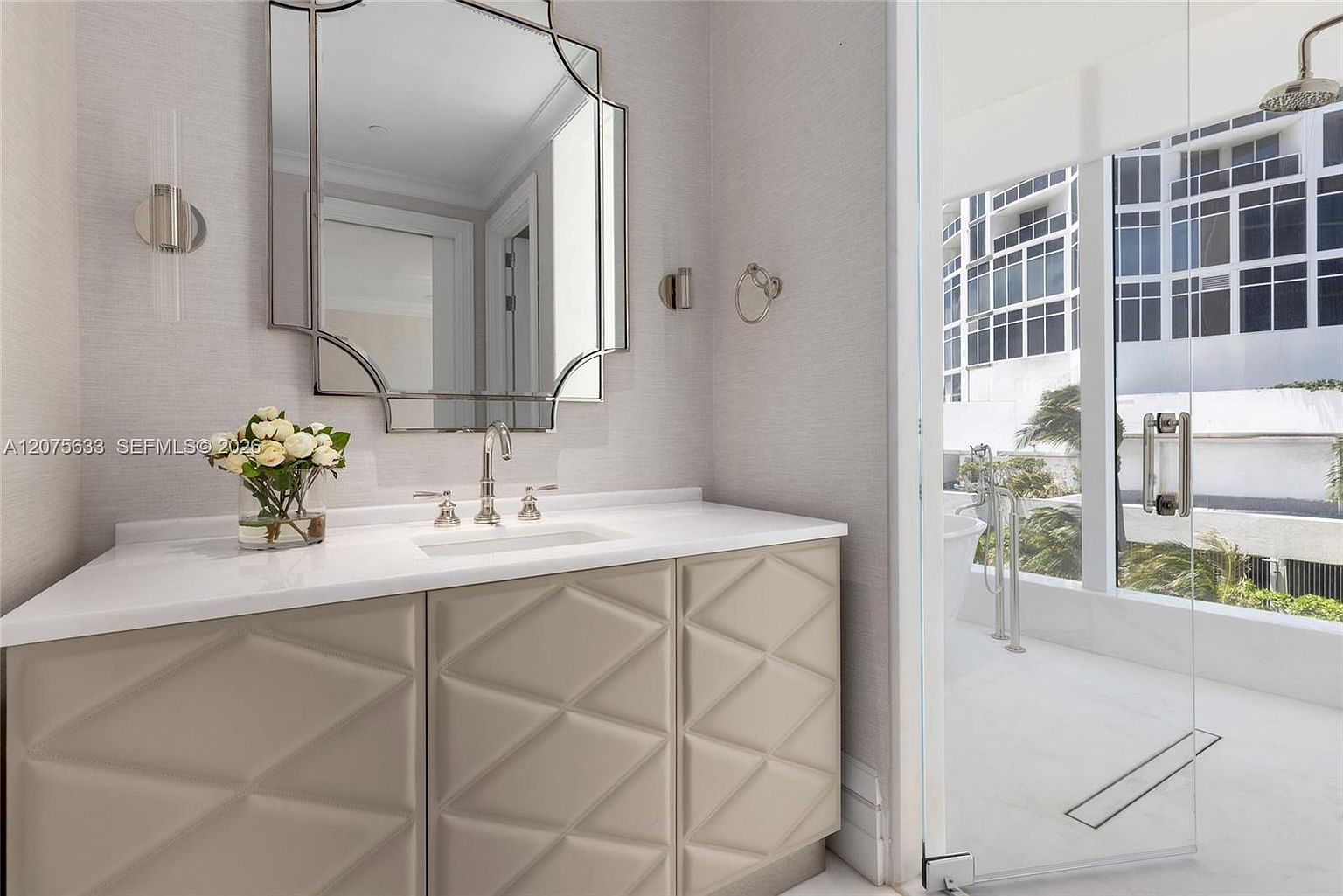 This elegant bathroom features a vanity with a textured, geometric-patterned cabinet base and a crisp white countertop. A decorative, multi-faceted mirror hangs above the sink, while a glass-enclosed shower area reveals a bright, airy space with a view of the outdoors. The neutral color palette and clean lines create a sophisticated and modern atmosphere.