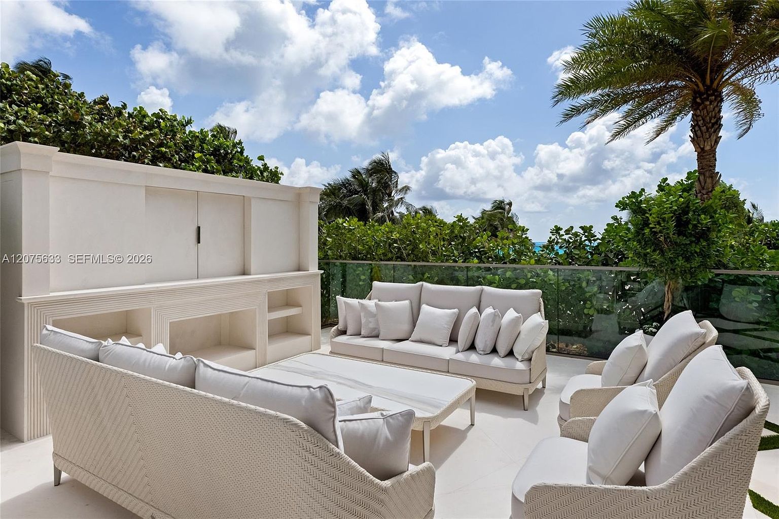 This elegant outdoor terrace features a sophisticated lounge area with light-toned wicker furniture and plush white cushions. A custom-built, light-colored storage unit with a central cabinet serves as a focal point, while a glass railing provides an unobstructed view of the lush greenery and blue sky beyond. The space is designed for relaxation, offering a serene atmosphere with a palm tree adding a tropical touch to the balcony setting.
