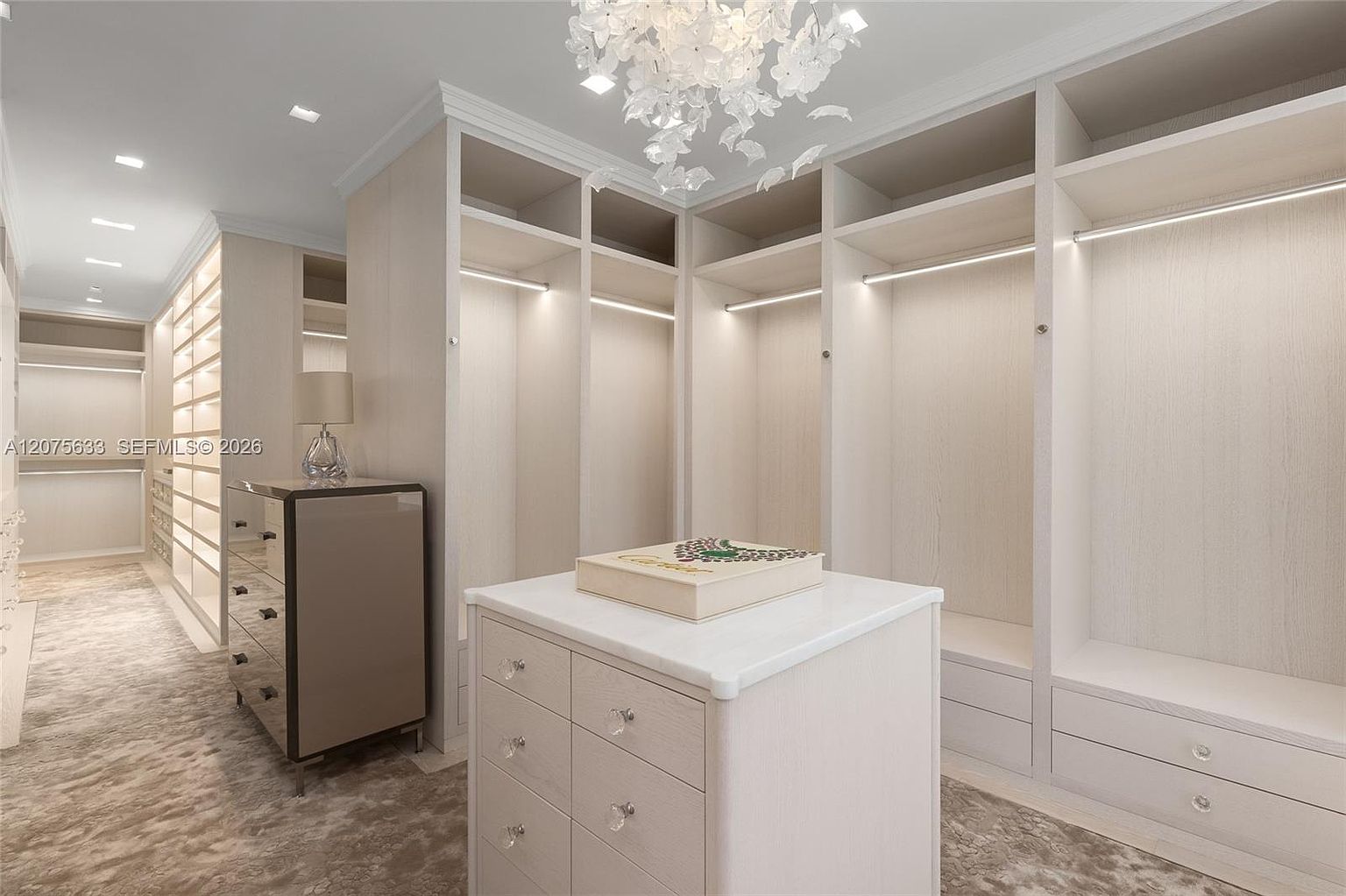 This luxurious walk-in closet features custom light-toned wood cabinetry with integrated LED lighting and a central island topped with a smooth, light-colored surface. A decorative floral-inspired chandelier hangs from the ceiling, adding a touch of elegance to the space, while a mirrored dresser sits to the left. The room is finished with plush carpeting and offers an expansive, organized storage solution.