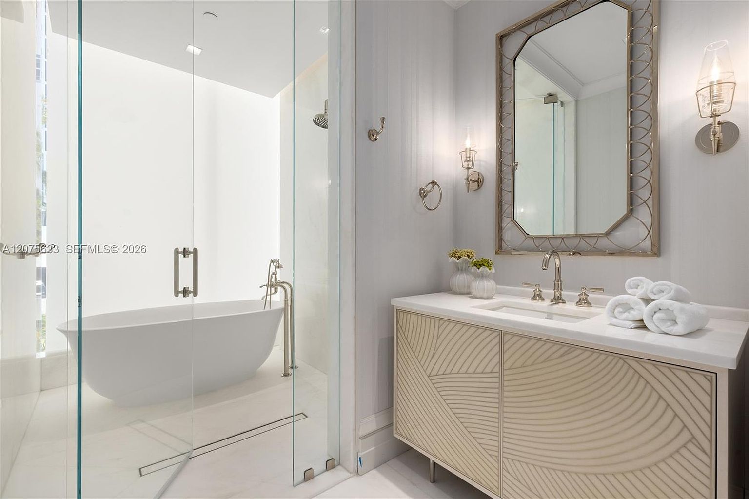 This elegant bathroom features a sleek, freestanding soaking tub enclosed behind a glass partition, creating a spa-like atmosphere. To the right, a vanity with a unique, textured geometric pattern on the cabinet doors is topped with a clean white counter and a decorative mirror. The space is illuminated by sophisticated wall sconces, contributing to a bright, modern, and luxurious aesthetic.
