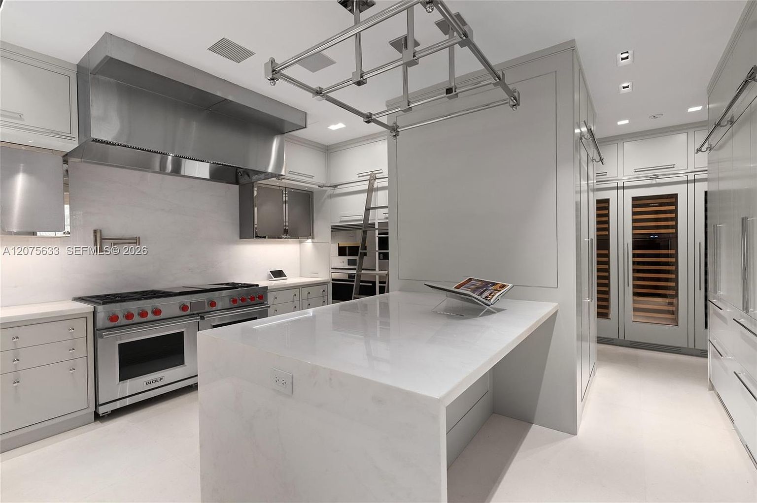 This sleek, modern kitchen features a minimalist aesthetic with light-toned cabinetry and a large central island topped with a smooth stone surface. A professional-grade stainless steel range with red knobs serves as a focal point beneath a substantial matching range hood, while a rolling library-style ladder provides access to upper storage. The space is illuminated by recessed lighting and a unique overhead metal rack, creating a sophisticated and highly functional culinary environment.