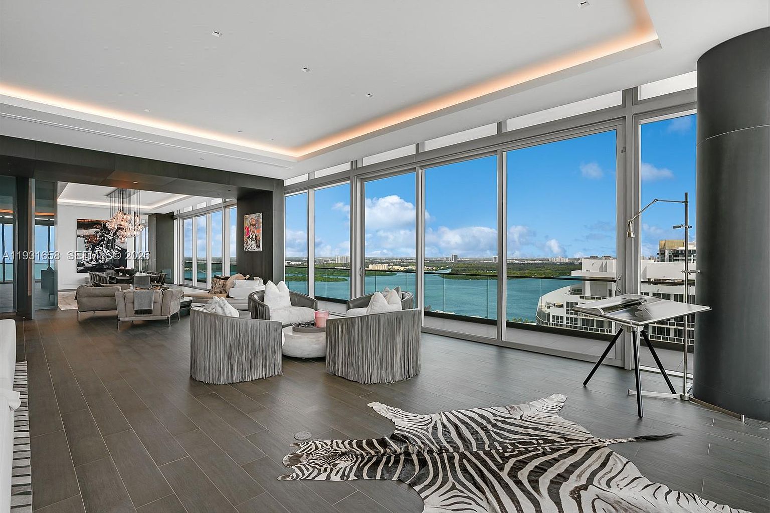 This is a luxurious living room with floor-to-ceiling windows offering a stunning water view. The room features modern furniture, including stylish armchairs and a zebra-print rug, complemented by dark wood flooring. The space is well-lit with recessed lighting and natural light, creating an elegant and inviting atmosphere.