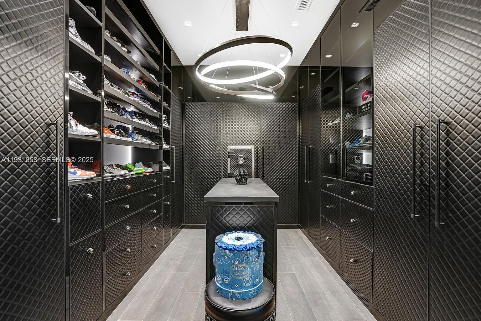This is a luxurious walk-in closet featuring custom shelving and drawers for shoe and clothing storage. The closet is finished with quilted black panels and illuminated by a modern circular chandelier. A central island with a decorative cake adds a unique touch to the space.