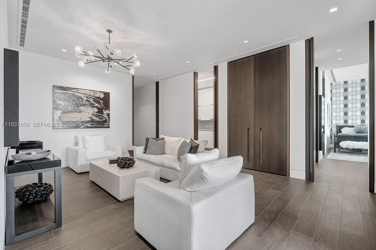 This is a bright and modern living room featuring white sofas and armchairs, a light-colored coffee table, and a striking abstract painting. The room is illuminated by a contemporary chandelier, and the hardwood flooring adds warmth. The perspective is from a medium angle, showcasing the layout and design elements.