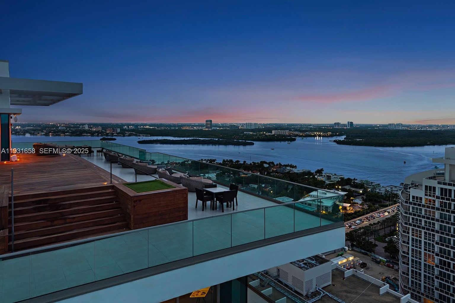 This image showcases a luxurious rooftop patio or balcony with modern design elements. The space features sleek glass railings, comfortable outdoor seating arrangements, and a wooden deck area. The view overlooks a stunning cityscape and waterfront at dusk, creating an upscale and inviting atmosphere.