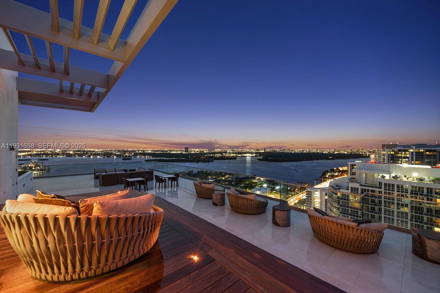 This image showcases a luxurious rooftop patio at dusk, featuring stylish wicker seating arrangements and a glass railing that offers panoramic views of the city skyline and waterfront. The deck is partially covered with wooden planks, adding warmth and texture to the modern design, creating an inviting outdoor living space perfect for relaxation and entertainment.