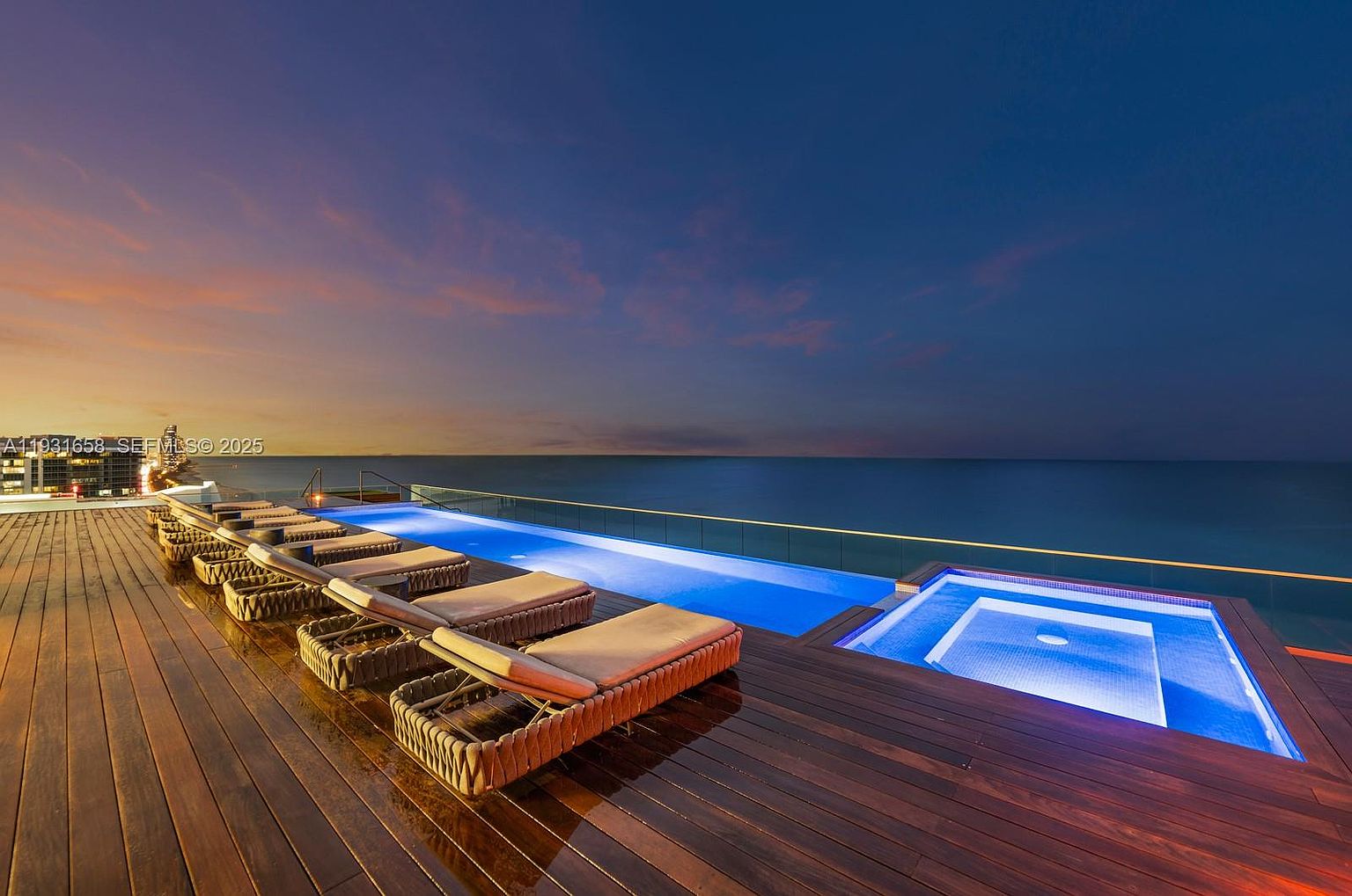 This image showcases a luxurious rooftop pool and spa area at dusk, featuring sleek lounge chairs arranged on a dark wooden deck. The infinity pool blends seamlessly with the ocean horizon, creating a serene and upscale ambiance. The spa is illuminated with blue light, adding to the modern and sophisticated feel of the property.