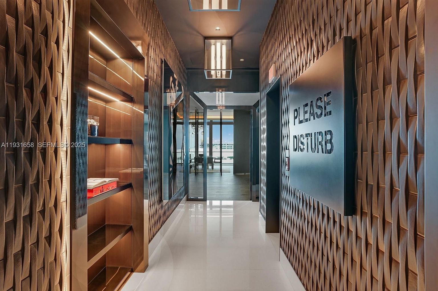 This is a modern hallway featuring textured walls, built-in shelving with accent lighting, and a 'Please Disturb' sign next to an elevator. The hallway has a sleek, contemporary design with glossy white floors and a view towards a bright, open space at the end. The perspective is a straight-on view down the hallway, emphasizing its length and design elements.