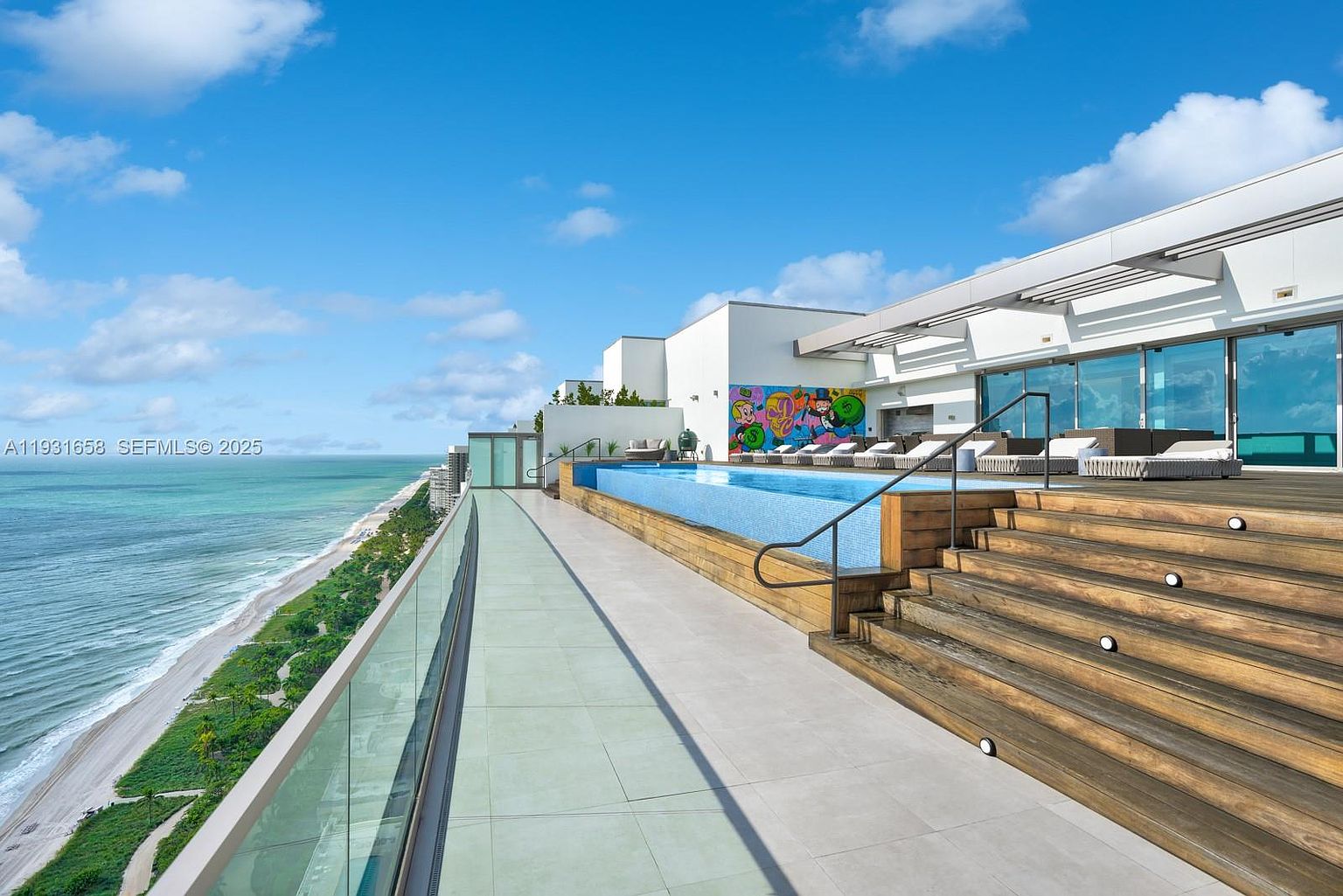 This image showcases a luxurious rooftop pool and deck area with stunning ocean views. The pool features a modern design with blue tiling, complemented by wooden decking and steps leading to lounge areas. A colorful mural adds a unique artistic touch, enhancing the upscale and relaxing atmosphere of this outdoor space.