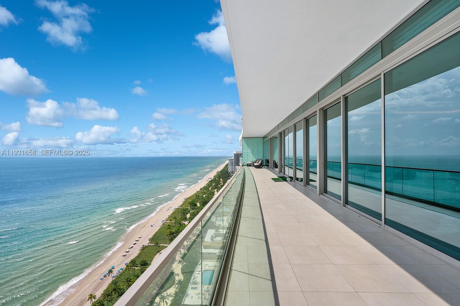 This image showcases a luxurious balcony with stunning ocean views. The balcony features a sleek glass railing, light-colored tile flooring, and floor-to-ceiling glass doors that seamlessly blend indoor and outdoor living. The overall impression is one of high-end coastal living with a focus on breathtaking scenery.