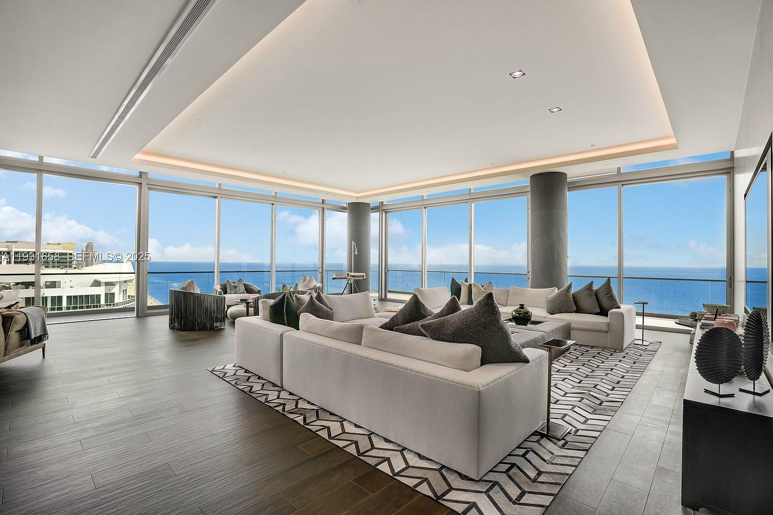 This is a luxurious living room with floor-to-ceiling windows offering panoramic ocean views. The room features a large, modern white sectional sofa with gray pillows, a geometric patterned rug, and contemporary decor. The open floor plan and abundant natural light create a spacious and inviting atmosphere.