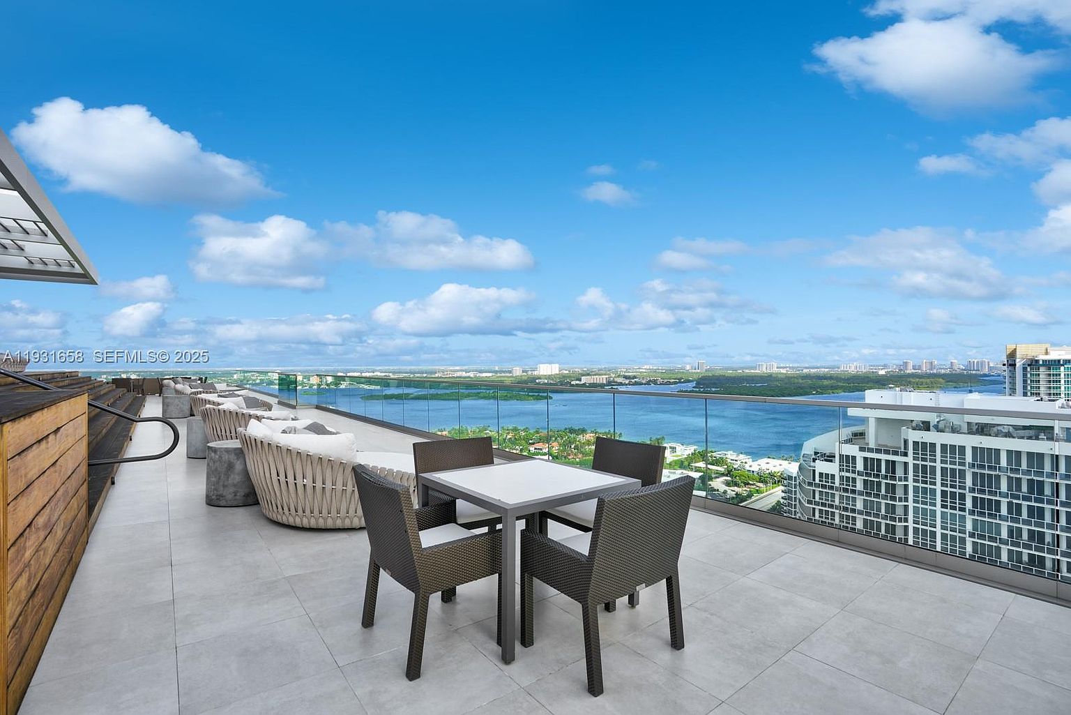 This image showcases a luxurious outdoor patio or balcony area with stunning panoramic views of the water and cityscape. The space is furnished with a modern dining set and comfortable lounge seating, suggesting an ideal setting for relaxation and entertainment. The clear glass railing enhances the unobstructed view, adding to the upscale ambiance.