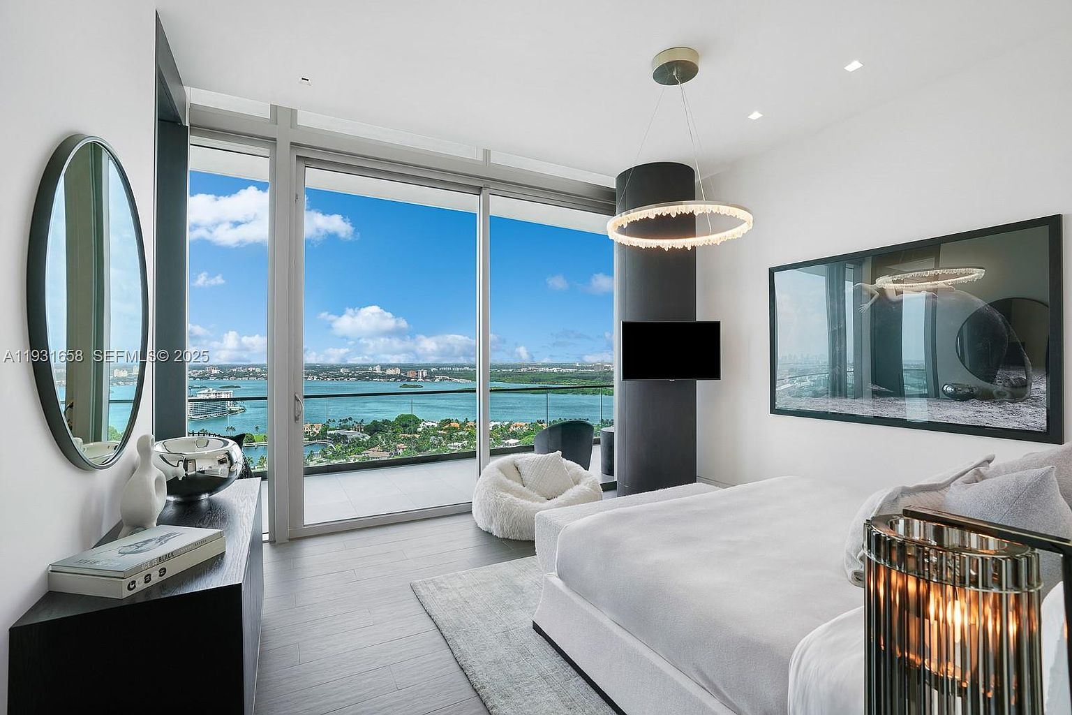 This is a luxurious primary bedroom featuring a modern design with a stunning view. The room includes a large bed, a stylish dresser with decorative items, and a comfortable beanbag chair. A large window offers panoramic views of the water and cityscape, enhancing the room's appeal and creating a serene atmosphere.