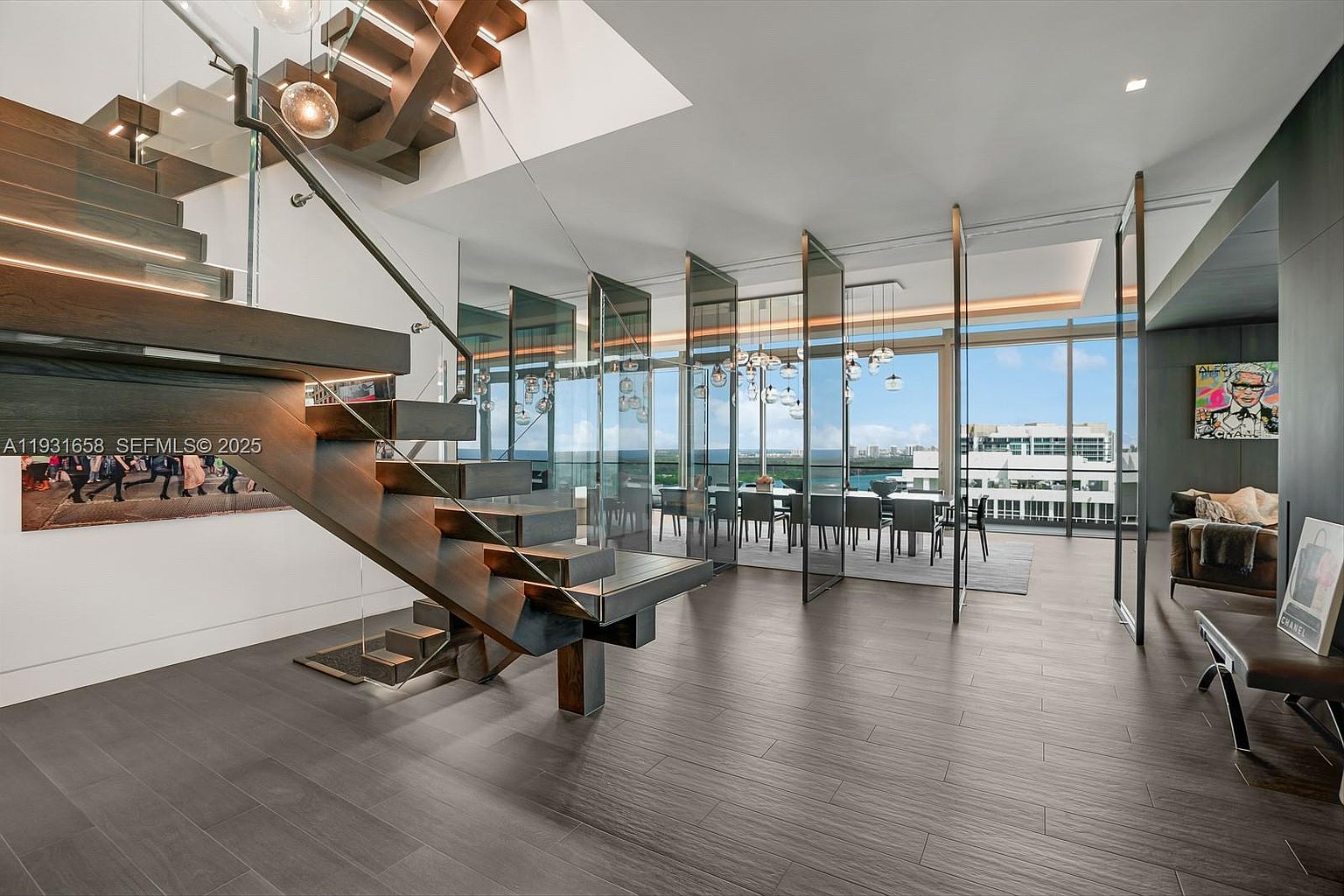 This interior shot showcases a modern home's entrance area, featuring a striking floating staircase with dark wood steps and glass railings. The open-concept design flows into a dining area with floor-to-ceiling windows offering expansive views, while contemporary artwork adds a touch of sophistication. The dark wood flooring complements the staircase and creates a cohesive, luxurious feel.