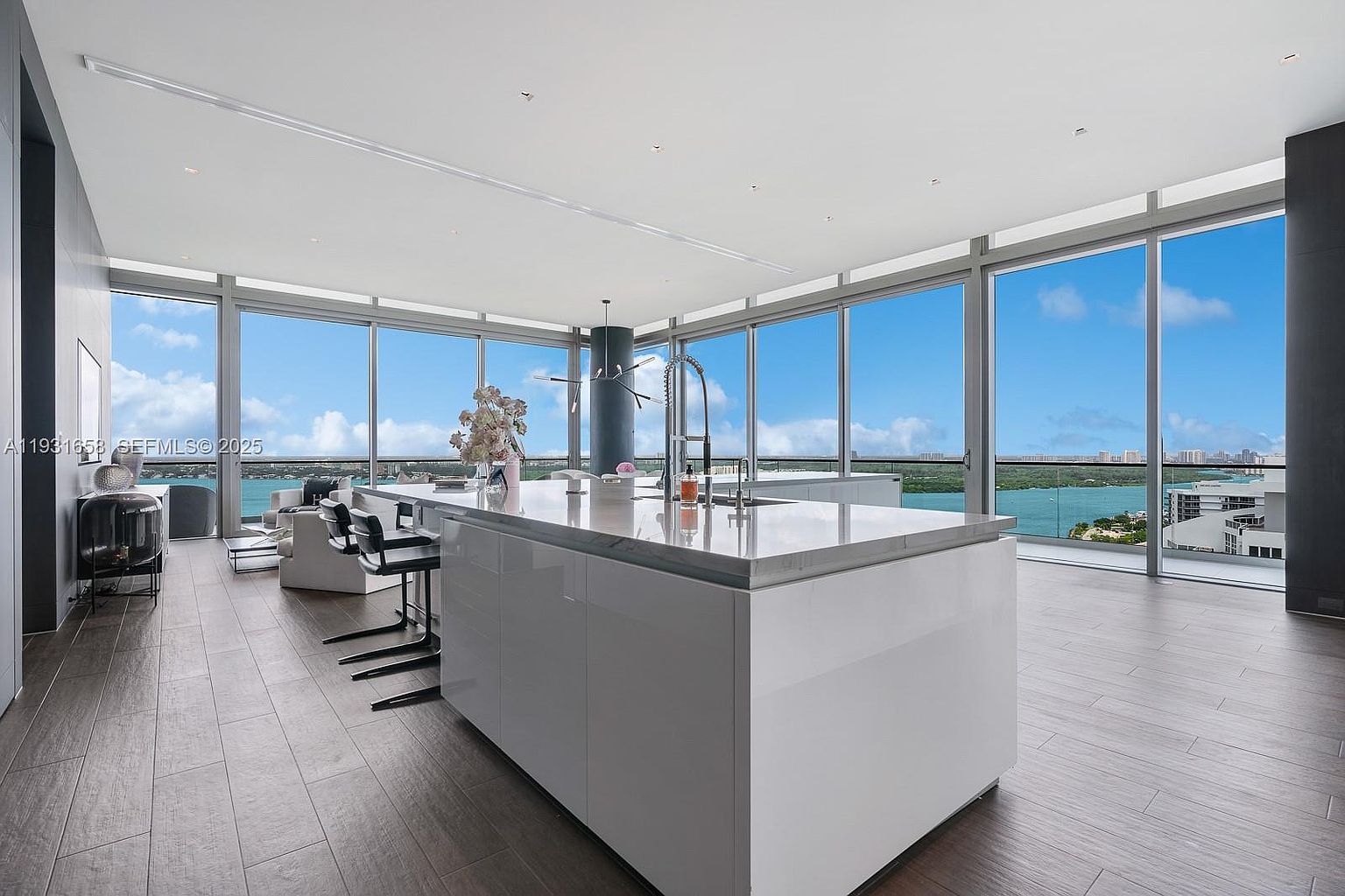 This interior shot showcases a modern, open-concept kitchen with a large white island as the focal point. Floor-to-ceiling windows offer expansive views of the water and cityscape, flooding the space with natural light. The kitchen features sleek cabinetry, modern bar stools, and a minimalist aesthetic, creating a luxurious and airy atmosphere.