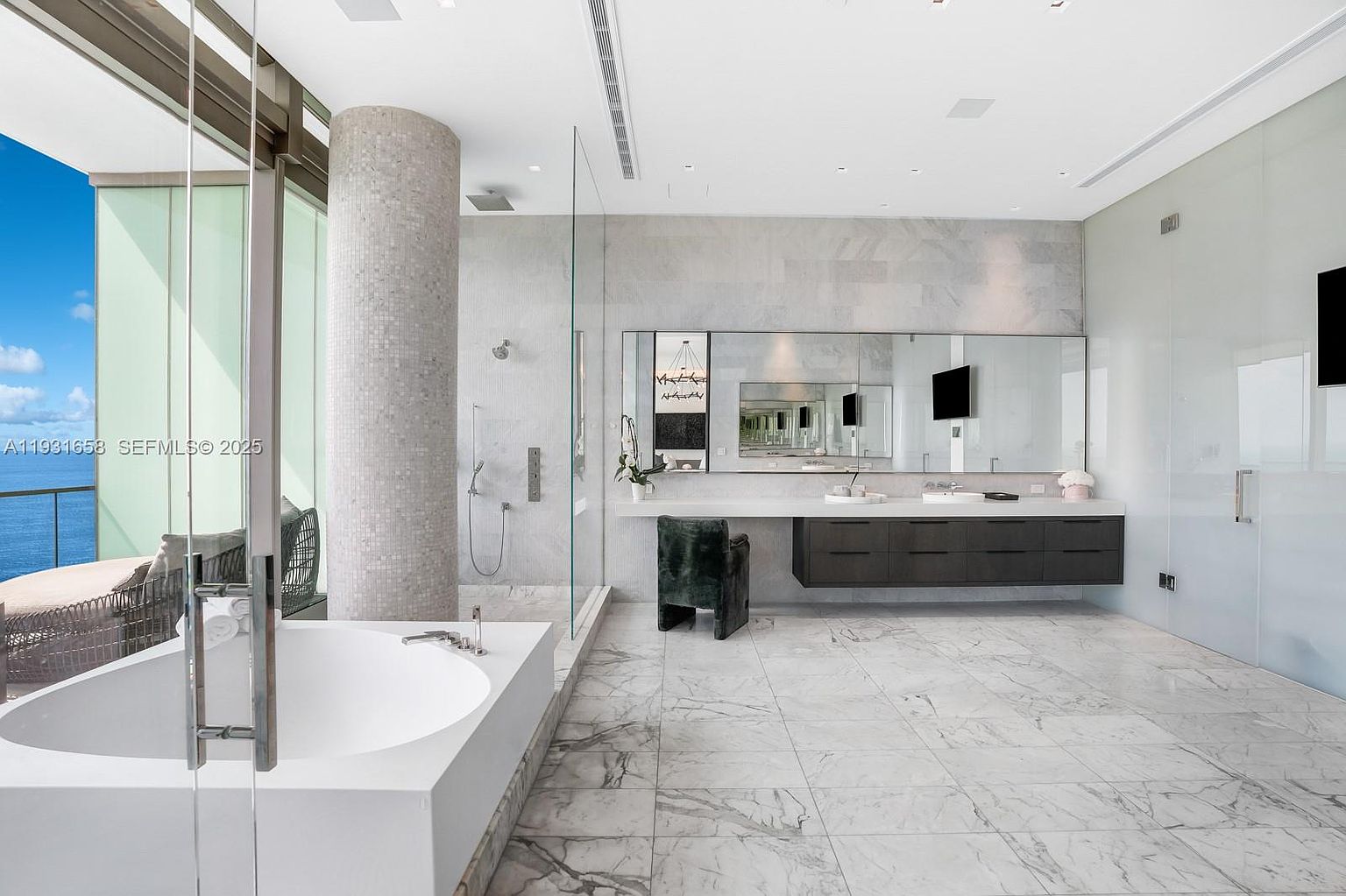 This is a luxurious primary bathroom featuring a modern freestanding bathtub next to a glass door leading to a balcony with ocean views. The bathroom boasts a spacious walk-in shower with a mosaic-tiled column, a floating vanity with a large mirror, and marble flooring. The overall impression is one of elegance and tranquility.