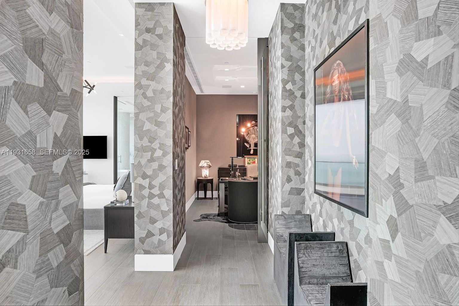 This interior shot showcases a modern hallway with unique geometric patterned wallpaper in shades of gray and white. The hallway leads to a bedroom and another room with a desk and artwork, illuminated by a contemporary chandelier. The flooring is light gray wood, and the overall impression is stylish and sophisticated.