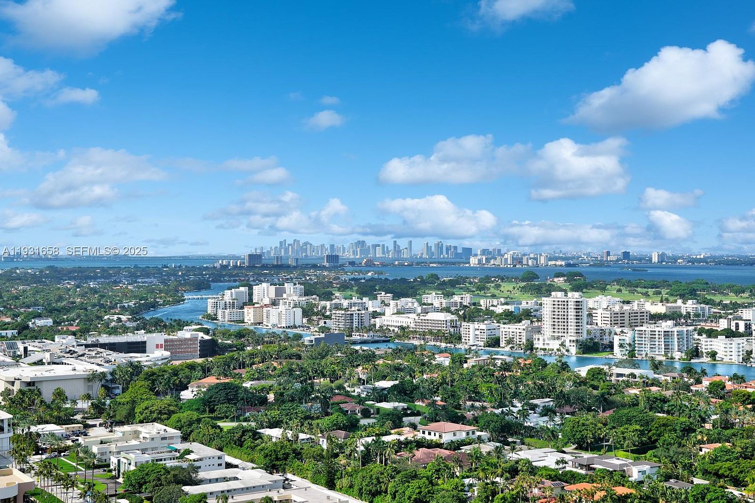 This aerial view showcases a sprawling cityscape with a mix of residential and commercial buildings, lush greenery, and waterways. The buildings are predominantly white, reflecting a modern architectural style. In the distance, a skyline of taller buildings is visible, suggesting a vibrant urban center, all under a bright blue sky dotted with fluffy white clouds.