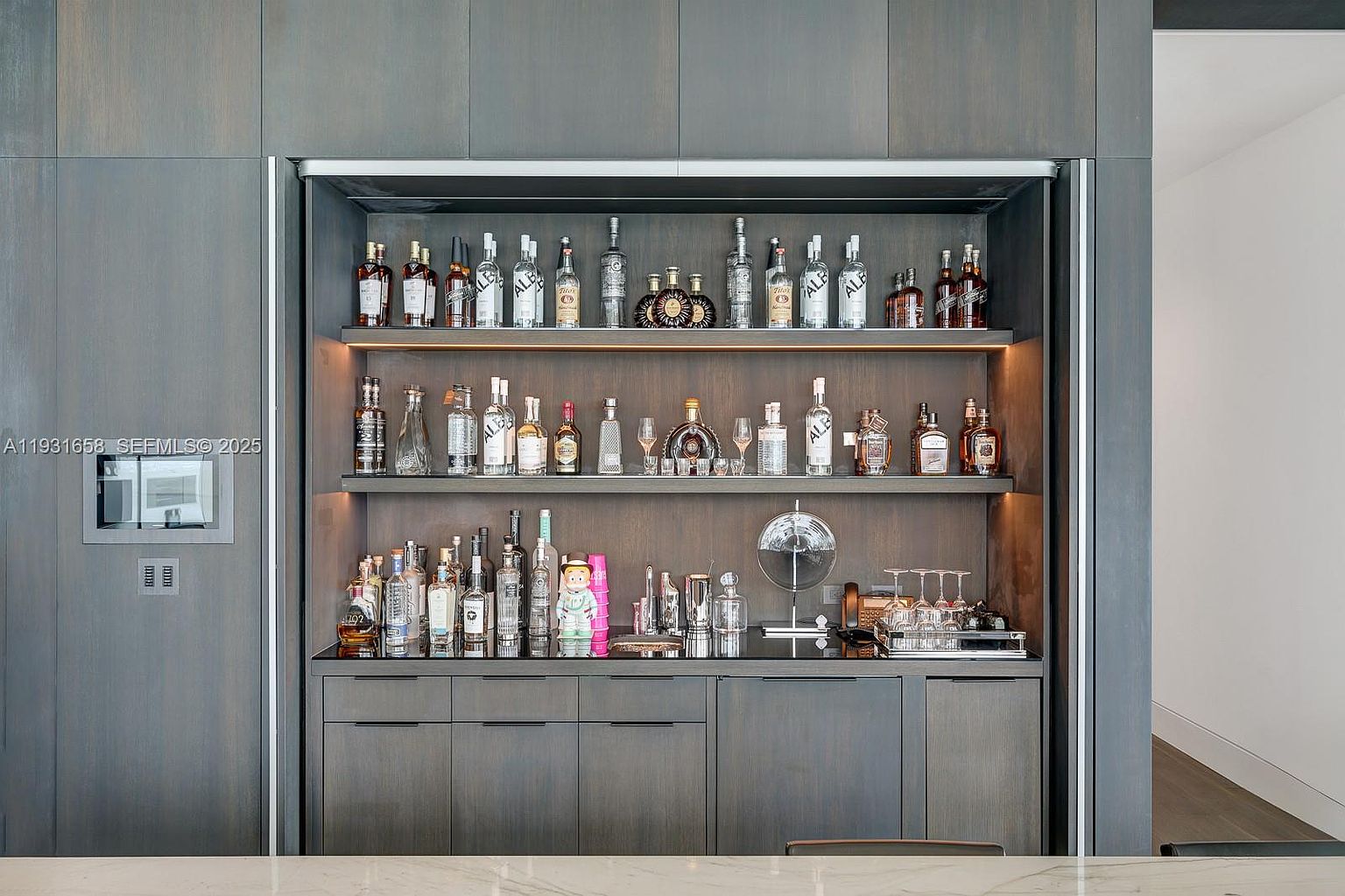 This image showcases a stylish built-in bar area, featuring dark wood cabinetry with sleek, minimalist hardware. The bar is well-stocked with various bottles and glassware, illuminated by subtle lighting that highlights the display. The overall design is modern and sophisticated, creating an inviting space for entertaining.