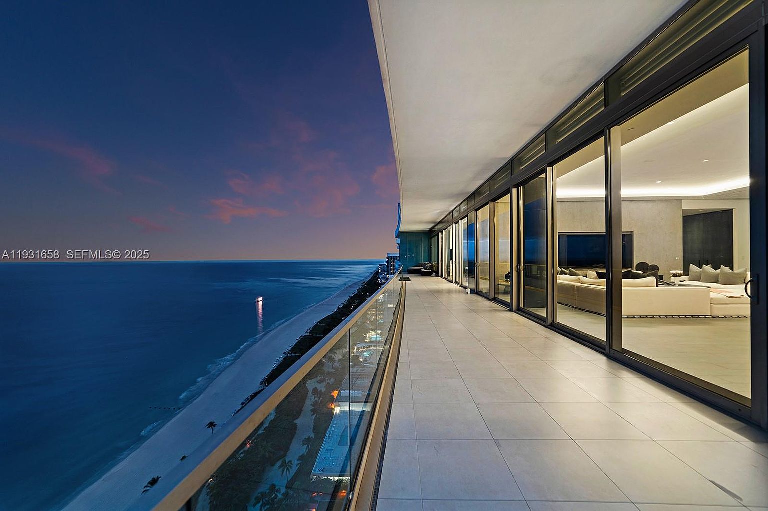 This image showcases a luxurious balcony with stunning ocean views at dusk. The balcony features sleek, modern design with glass railings and light-colored tile flooring. Sliding glass doors provide access to the interior, revealing a glimpse of a well-appointed living space with comfortable seating and ambient lighting, creating an inviting indoor-outdoor living experience.