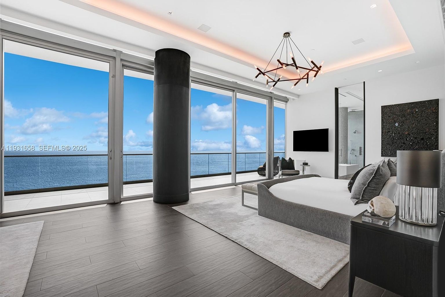 This is a luxurious primary bedroom with a modern design. The room features floor-to-ceiling windows offering stunning ocean views, a contemporary chandelier, and a sleek, low-profile bed. The dark wood flooring and neutral color palette create a sophisticated and relaxing atmosphere.