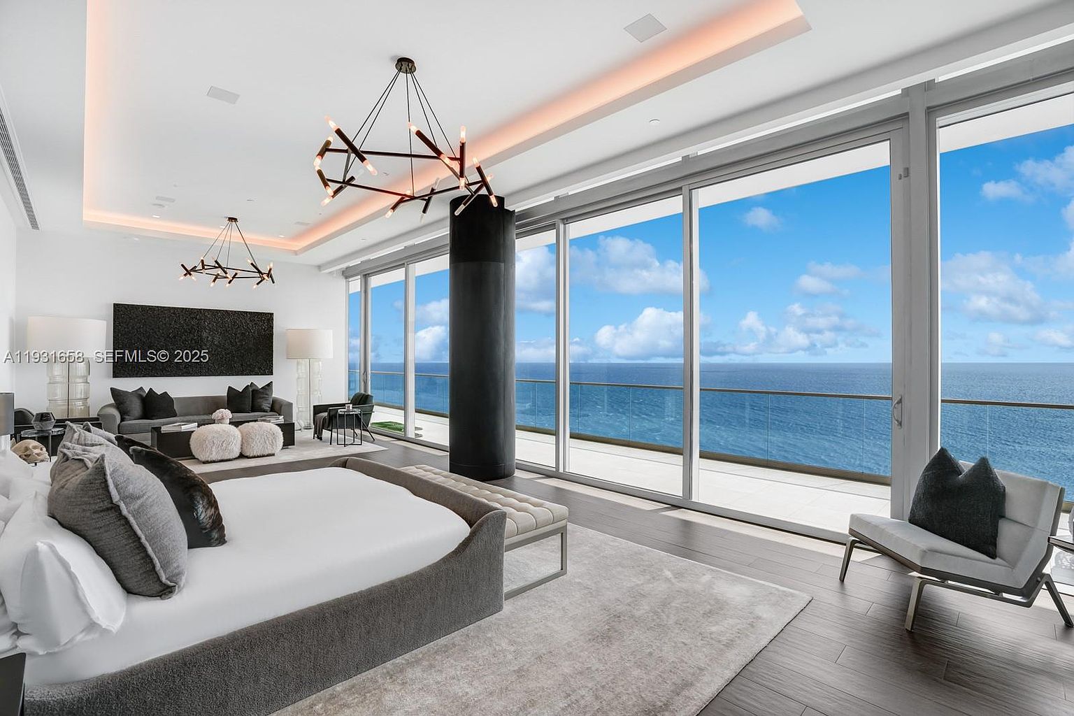 This is a luxurious primary bedroom with floor-to-ceiling windows offering stunning ocean views. The room features a modern design with a large bed, stylish lighting fixtures, and a comfortable seating area. The color palette is neutral, creating a serene and sophisticated atmosphere.