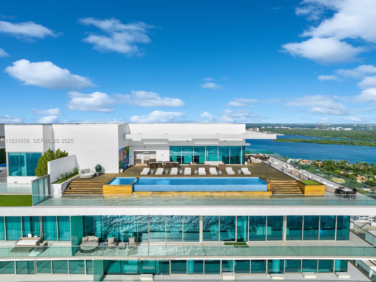 This aerial shot showcases a luxurious rooftop pool and deck area of a modern building. The pool is surrounded by lounge chairs and a wooden deck, with additional seating areas and a grill visible. The building features large glass windows, offering stunning views of the surrounding water and cityscape, creating an impression of high-end urban living.