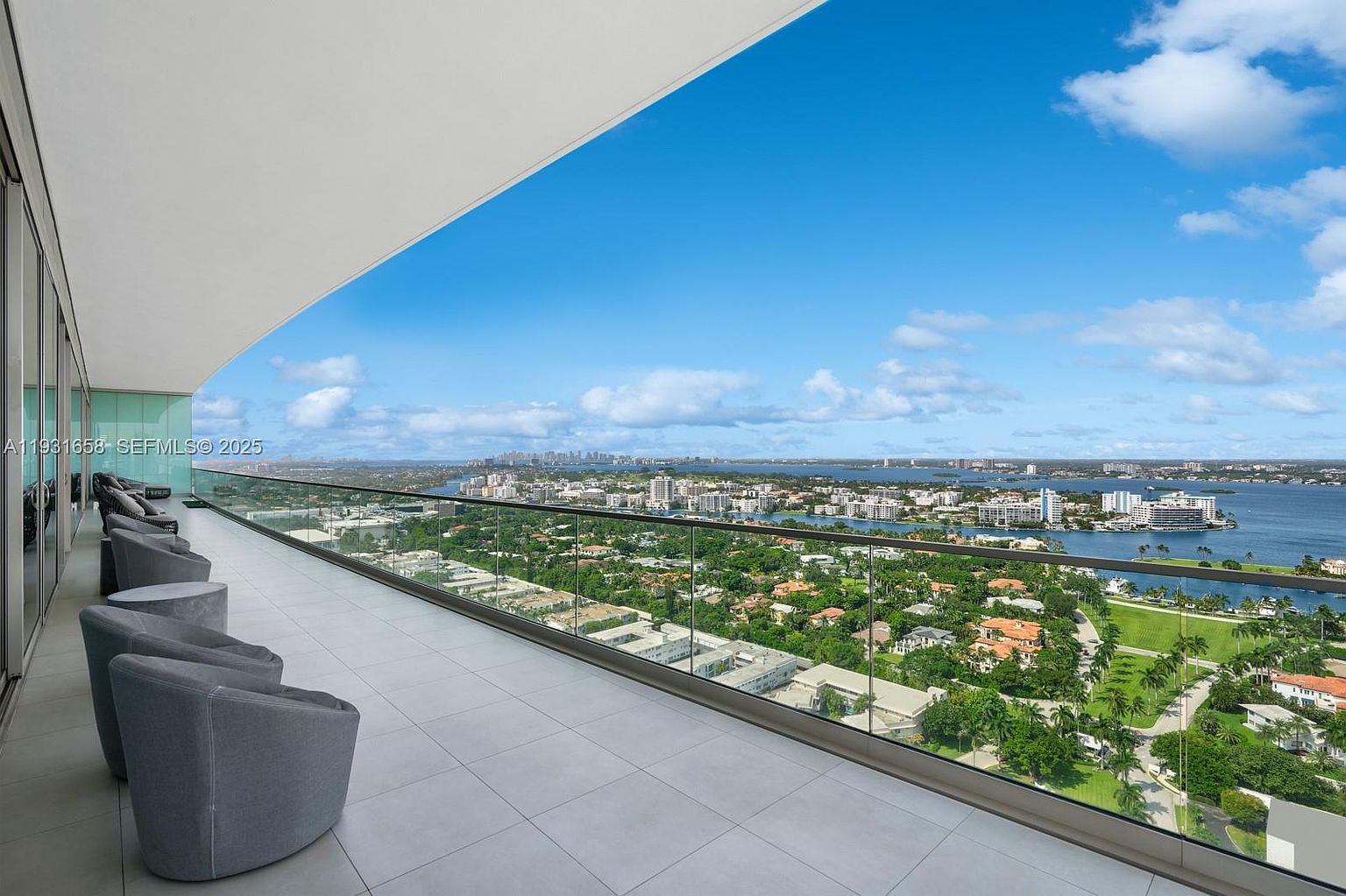 This image showcases a luxurious balcony with a stunning panoramic view of the city skyline and waterfront. The balcony features modern gray lounge chairs and a glass railing, providing an unobstructed view. The tiled floor adds a touch of elegance, making it an ideal space for relaxation and entertainment.