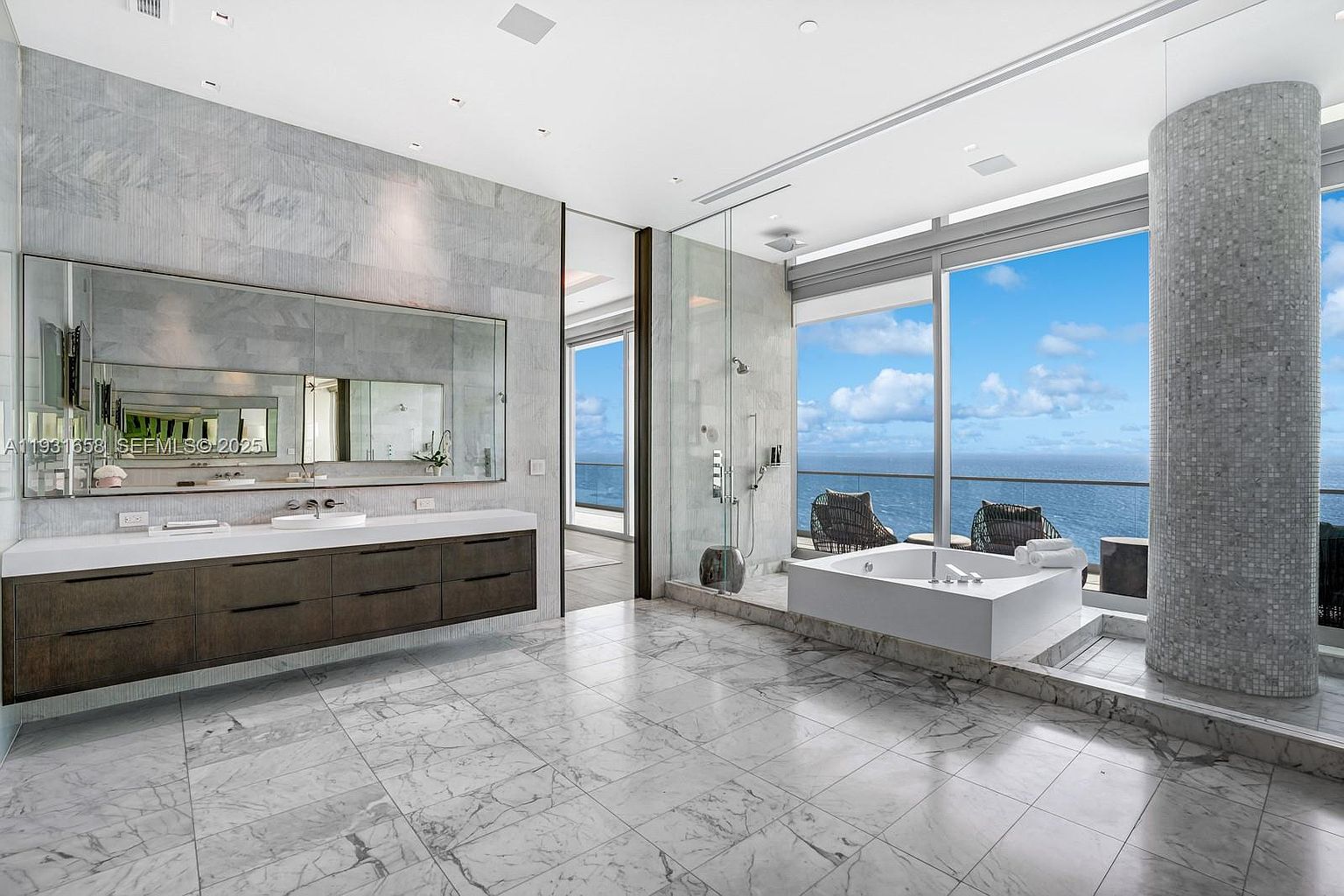 This is a luxurious primary bathroom featuring marble flooring and wall accents. A large vanity with dark wood cabinets and a white countertop is complemented by a wide mirror. The bathroom also includes a modern bathtub positioned near large windows offering ocean views, and a glass-enclosed shower area. The overall impression is one of spaciousness, elegance, and high-end design.