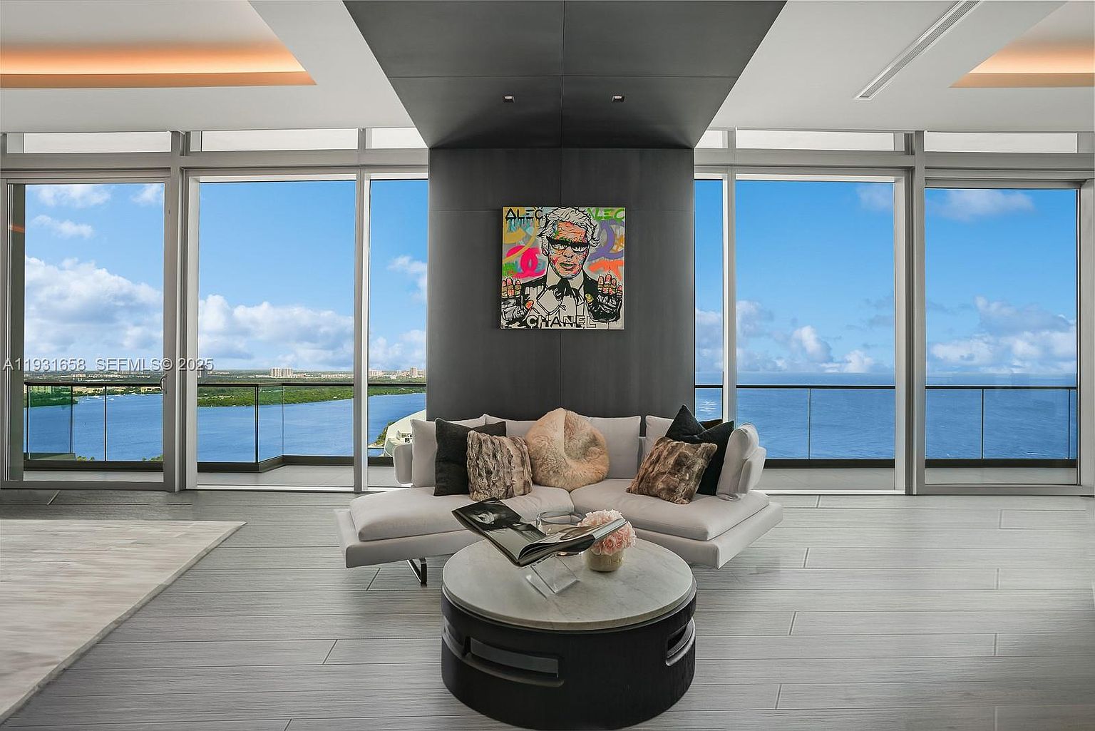 This is an interior shot of a modern living room with floor-to-ceiling windows offering a stunning ocean view. The room features a white sectional sofa with decorative pillows, a round coffee table with books and flowers, and a striking piece of pop art hanging above the sofa. The overall aesthetic is luxurious and contemporary, emphasizing the view and natural light.