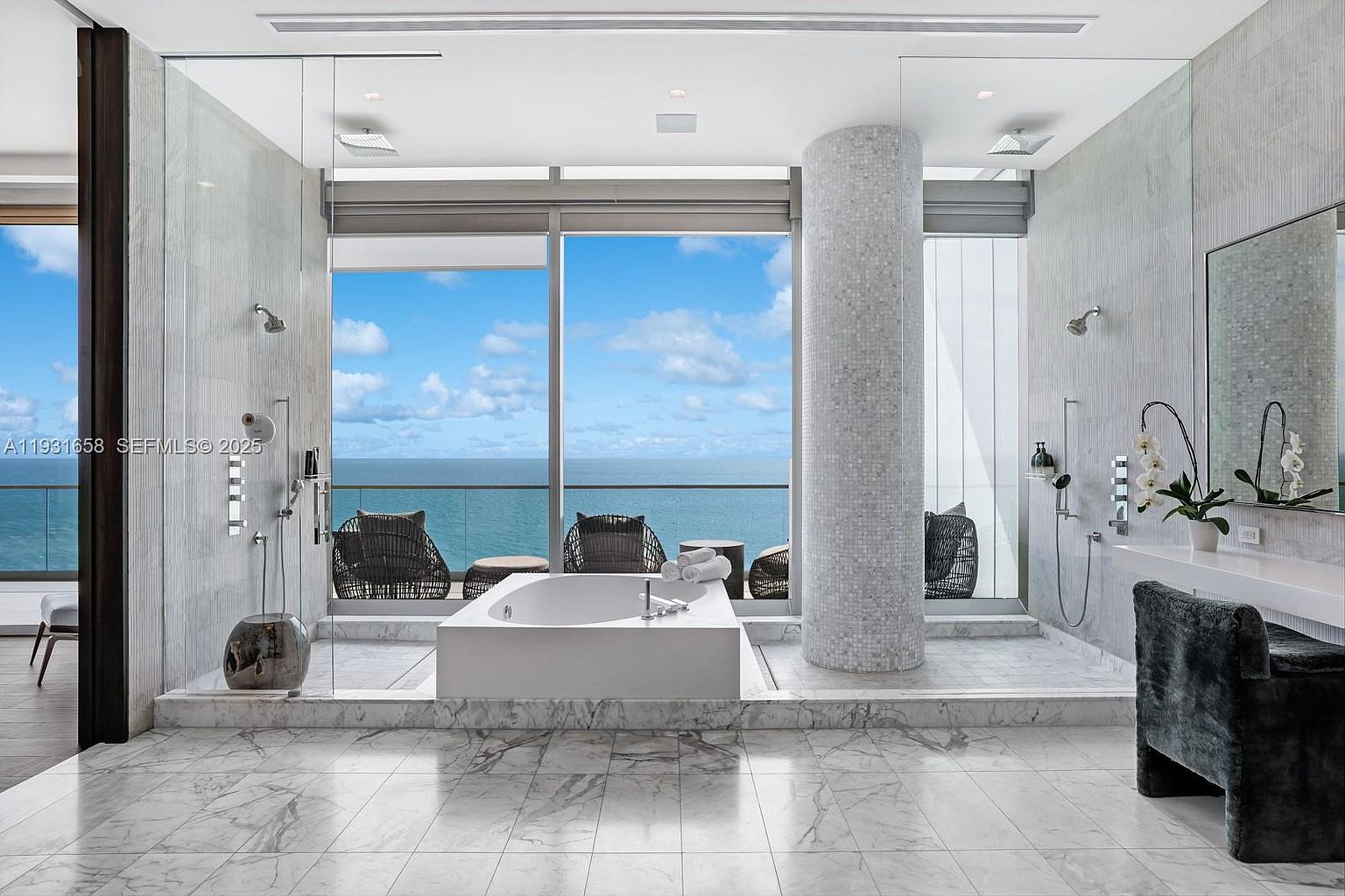 This is a luxurious primary bathroom featuring a freestanding bathtub with ocean views. The bathroom has a modern design with marble flooring and walls, a glass-enclosed shower, and a mosaic-tiled column. The space is bright and airy, creating a spa-like atmosphere.