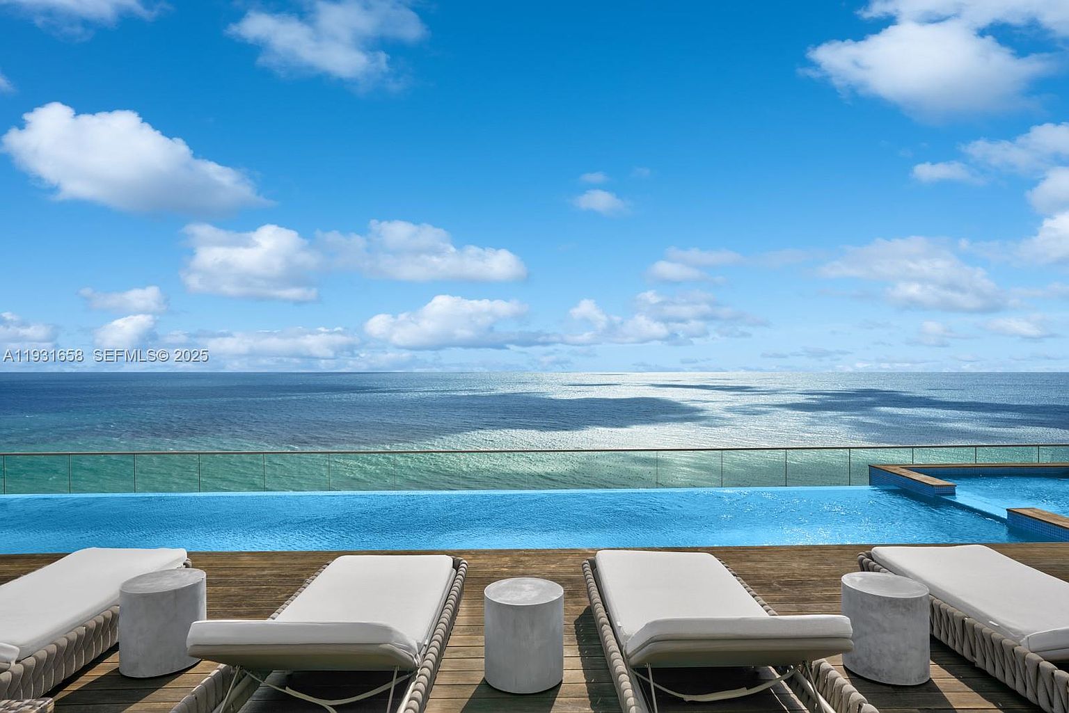 This image showcases a luxurious pool area with stunning ocean views. The pool is surrounded by a wooden deck furnished with modern lounge chairs and small side tables. The clear blue water of the pool blends seamlessly with the ocean horizon, creating a serene and inviting atmosphere.